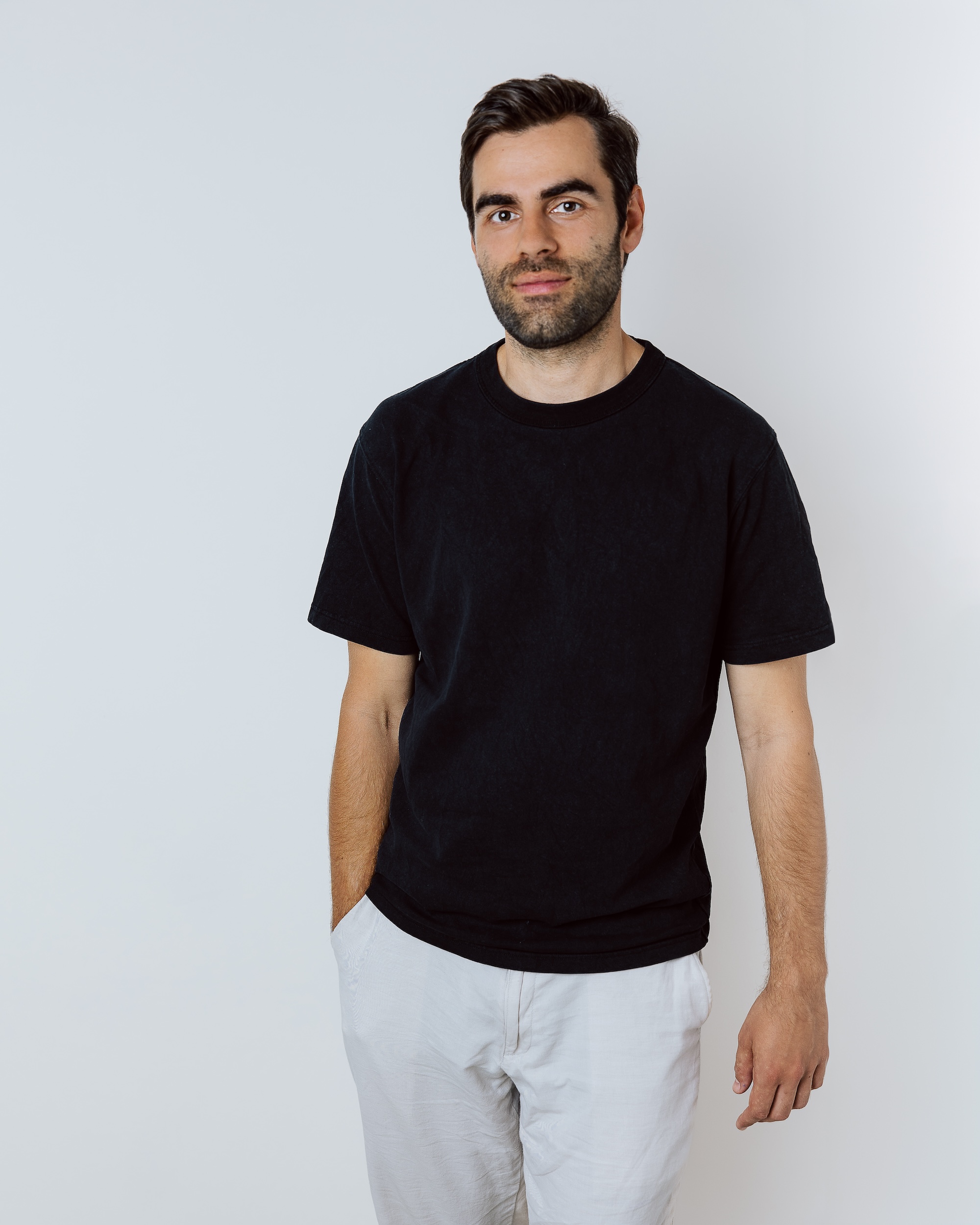 Man with beard wearing black t-shirt and white pants standing against a white background.