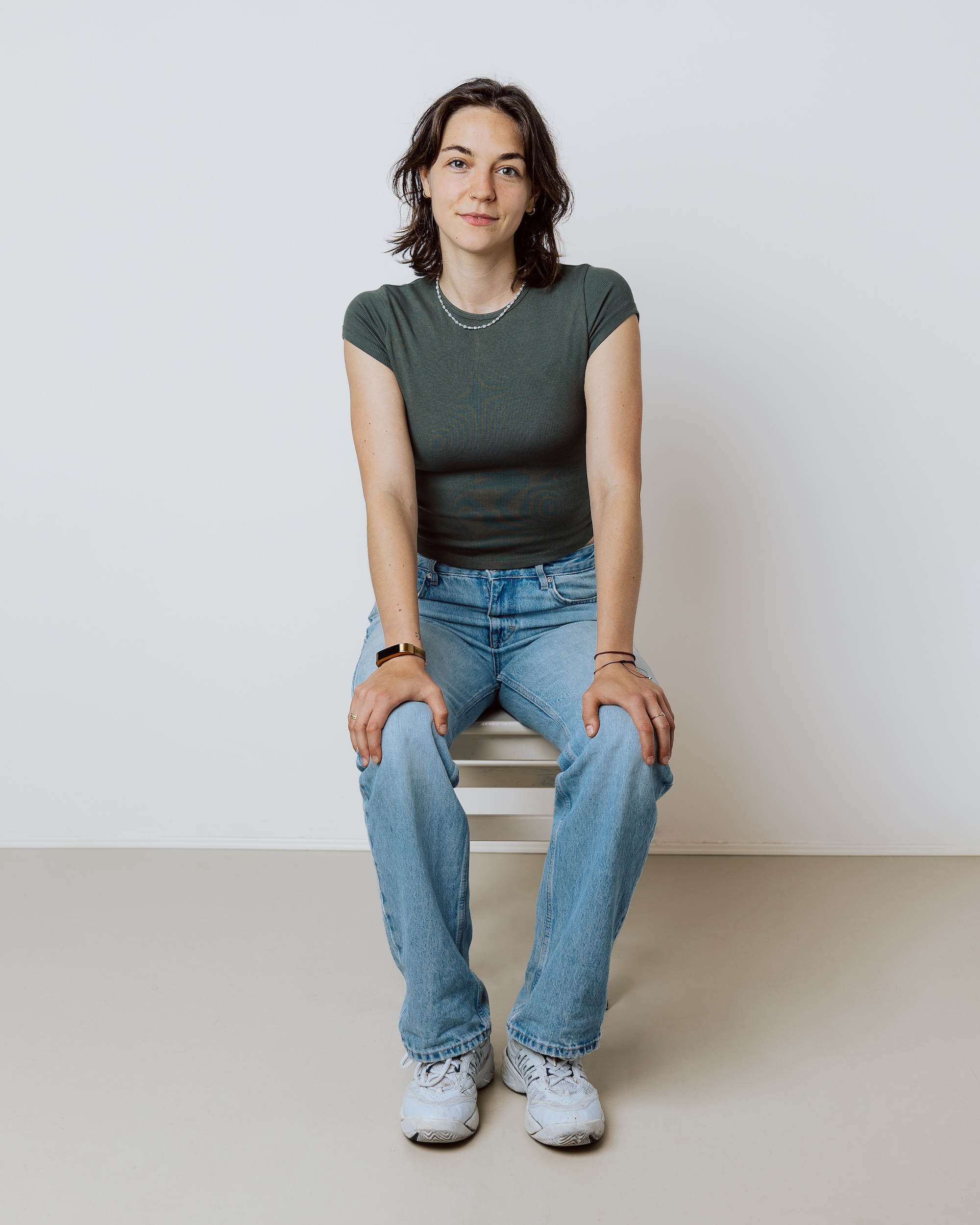 Young woman in green shirt and jeans sitting on a white chair against a plain background.
