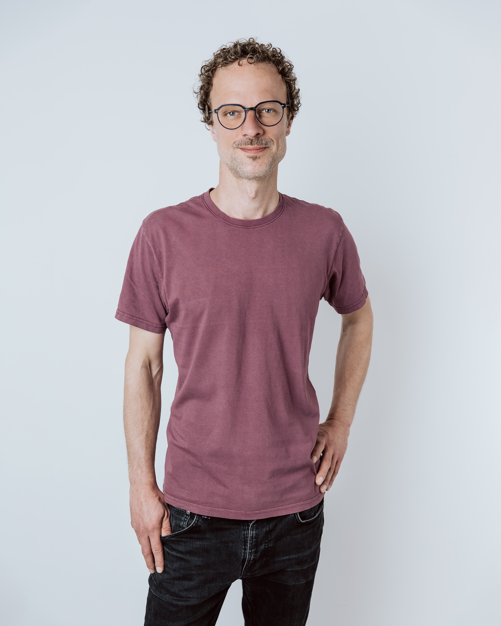 Man with curly hair and glasses wearing a maroon t-shirt and black jeans standing against a white wall.