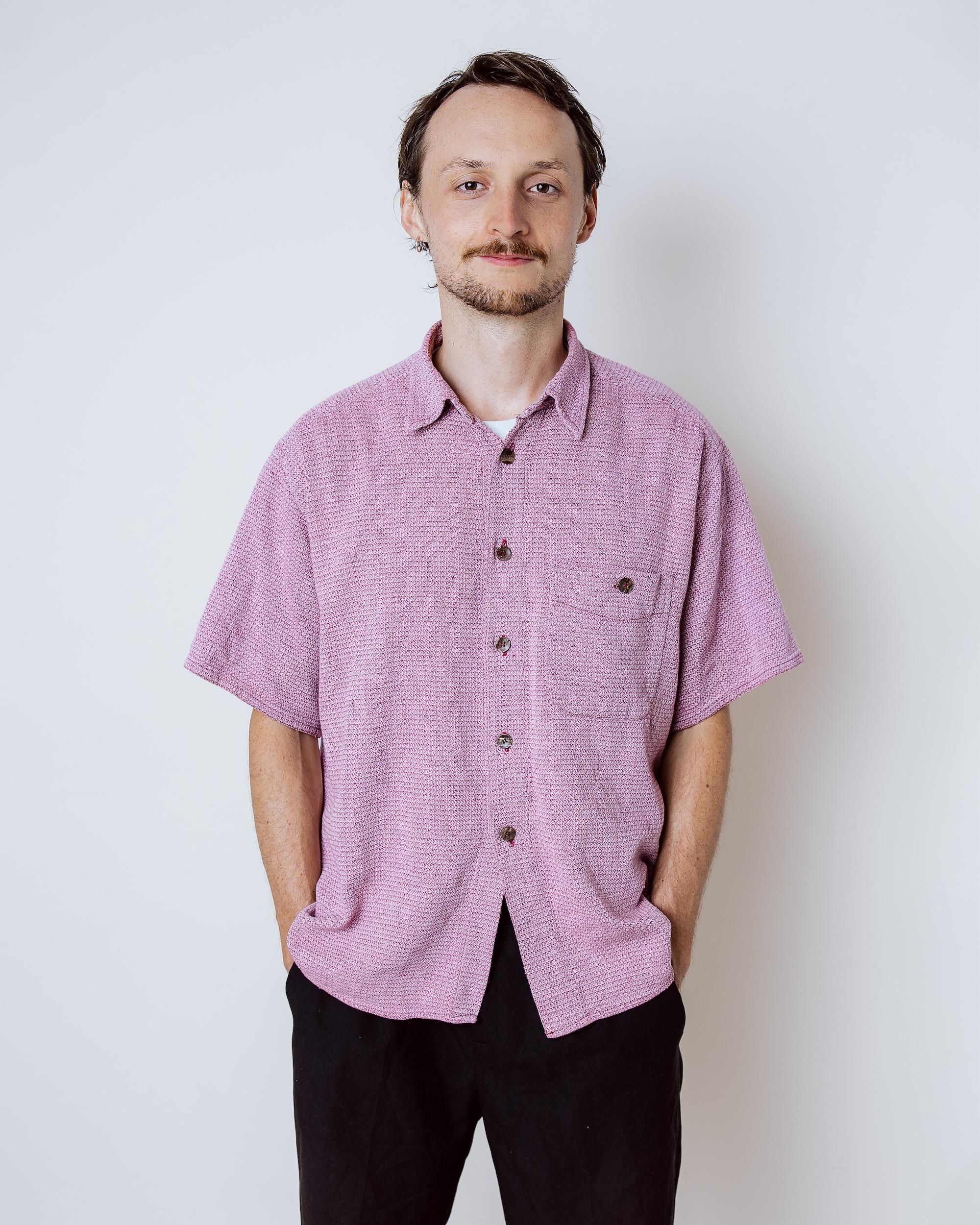 Man with short hair and beard wearing a pink button-up shirt and black pants, standing with hands in pockets.