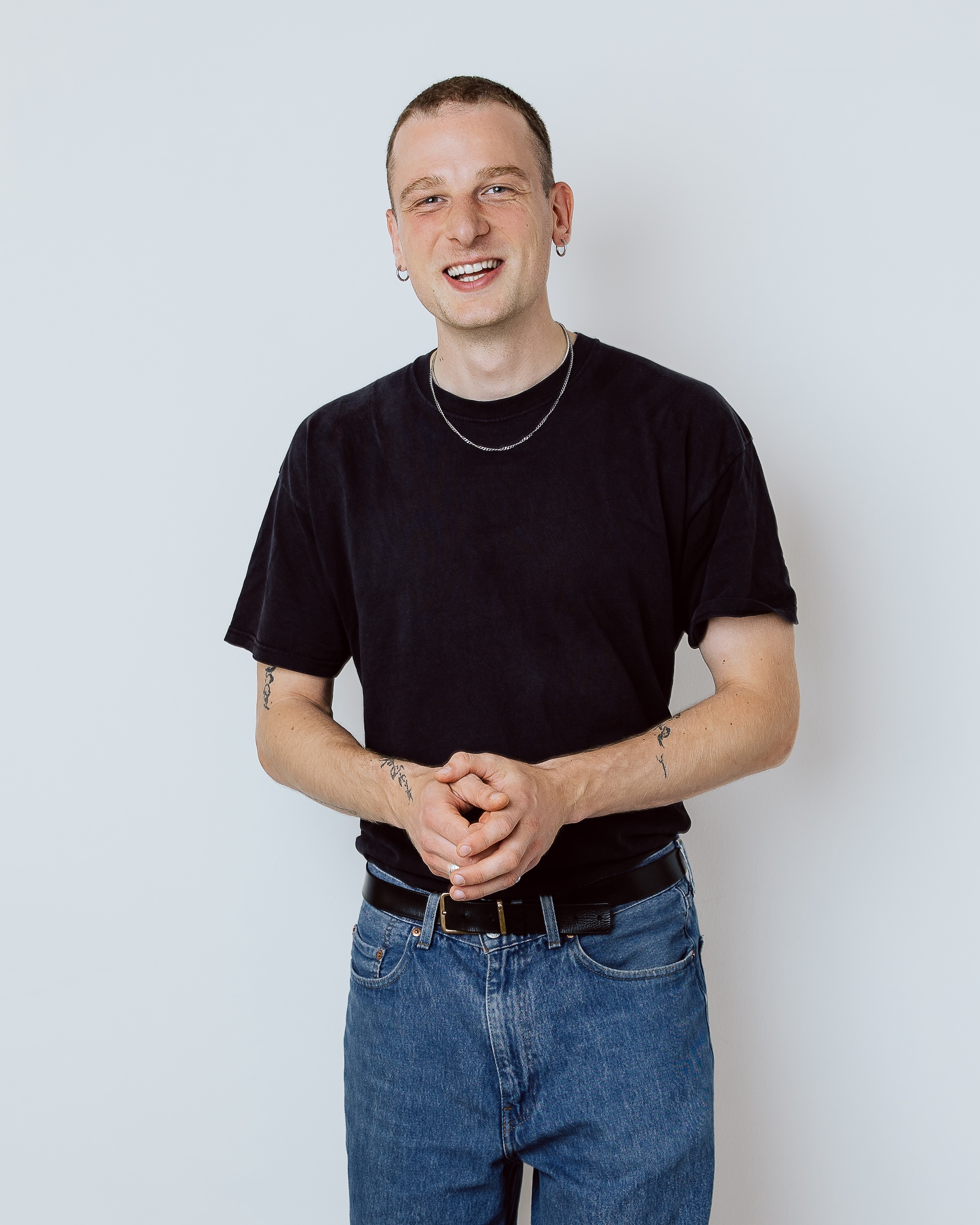 Smiling man in a black t-shirt and jeans stands with hands clasped against a white wall.