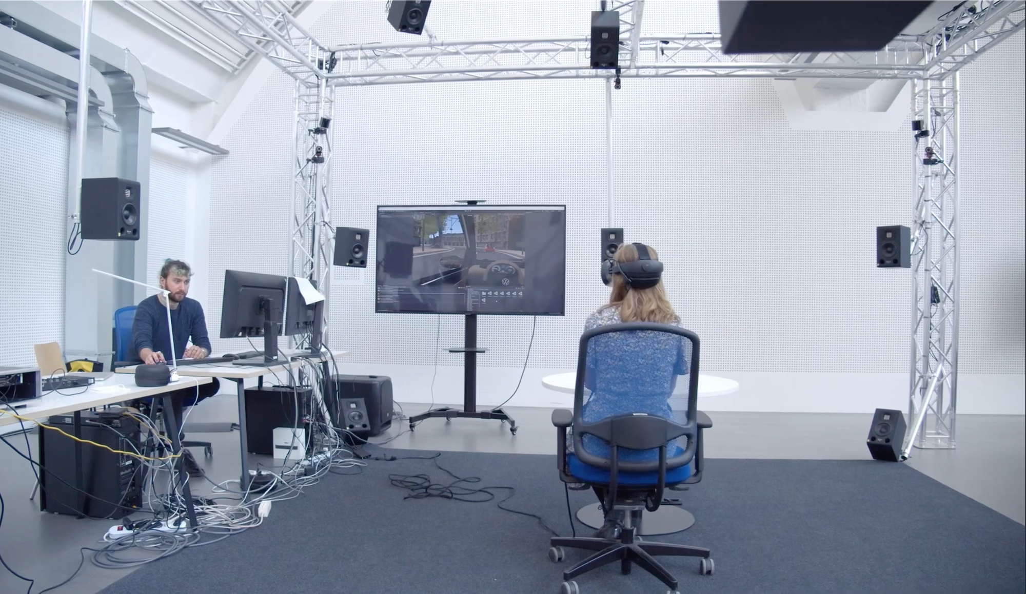 Person wearing VR headset facing screen with driving simulation, another person at desk with computer monitors.