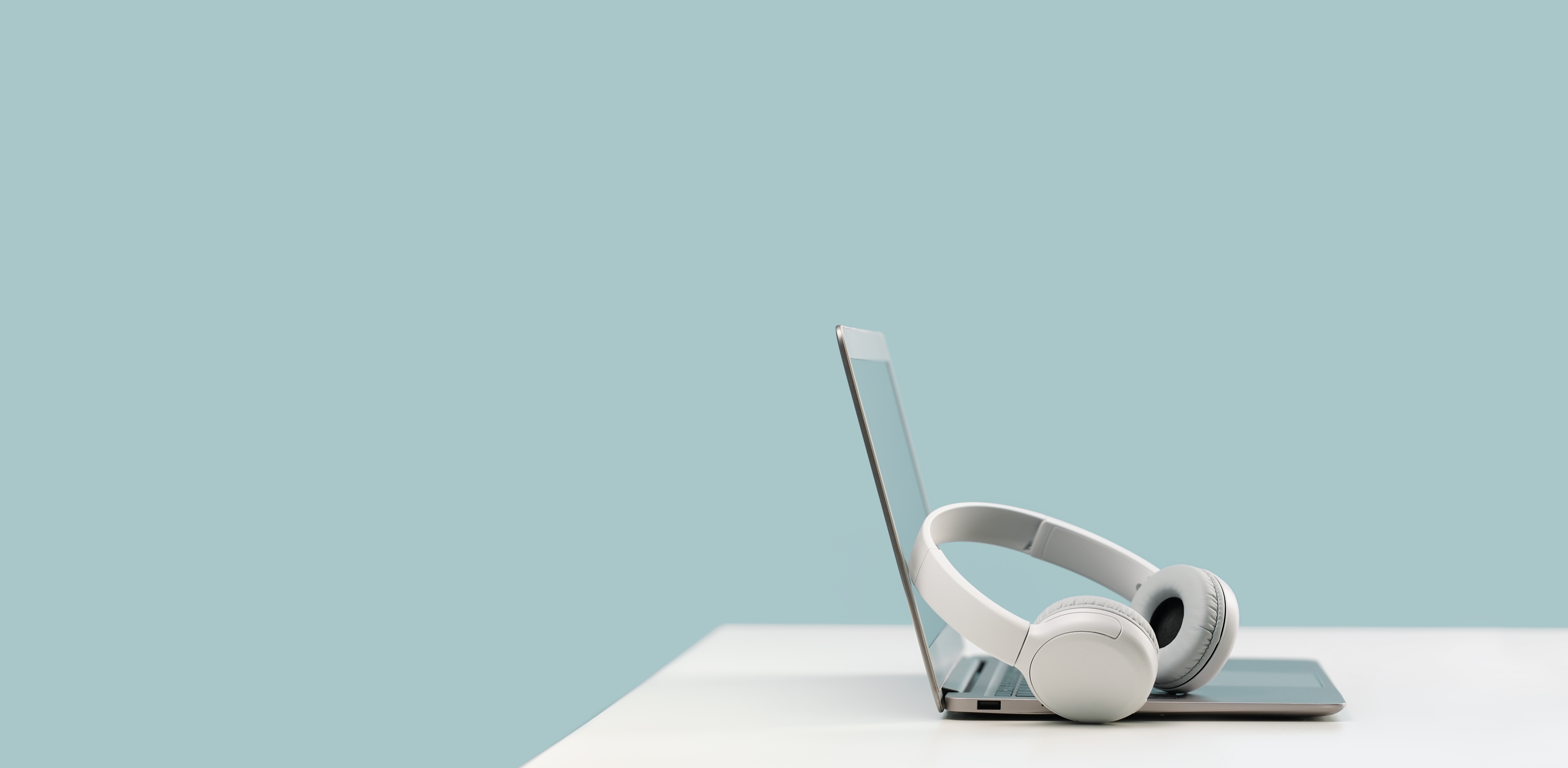White headphones resting on the keyboard of a slim, open laptop on a white surface with blue background.