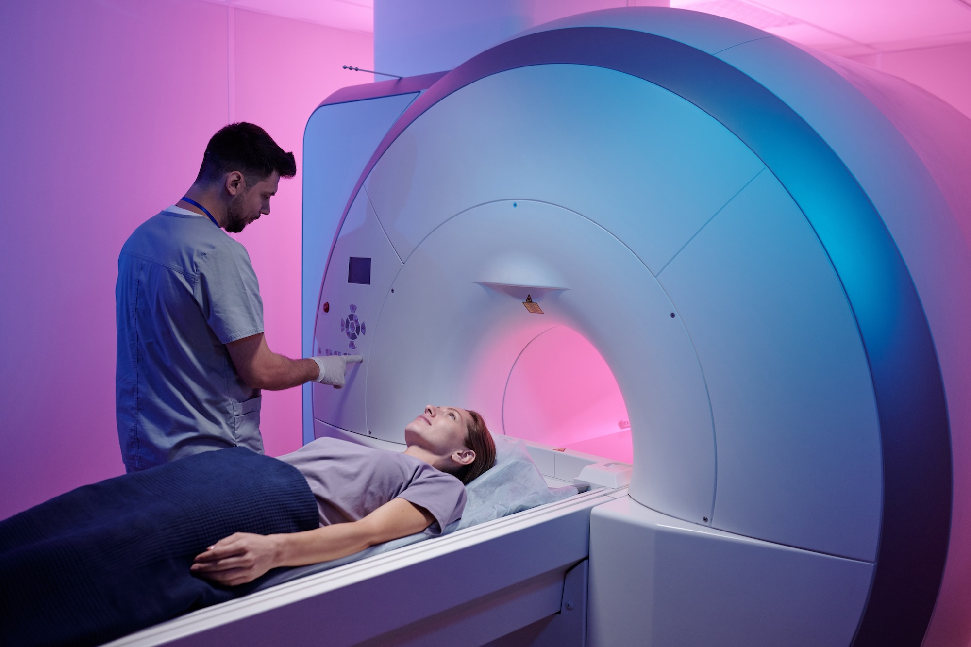 A male technician operates an MRI machine while a woman lies on the scanner bed under a blanket.