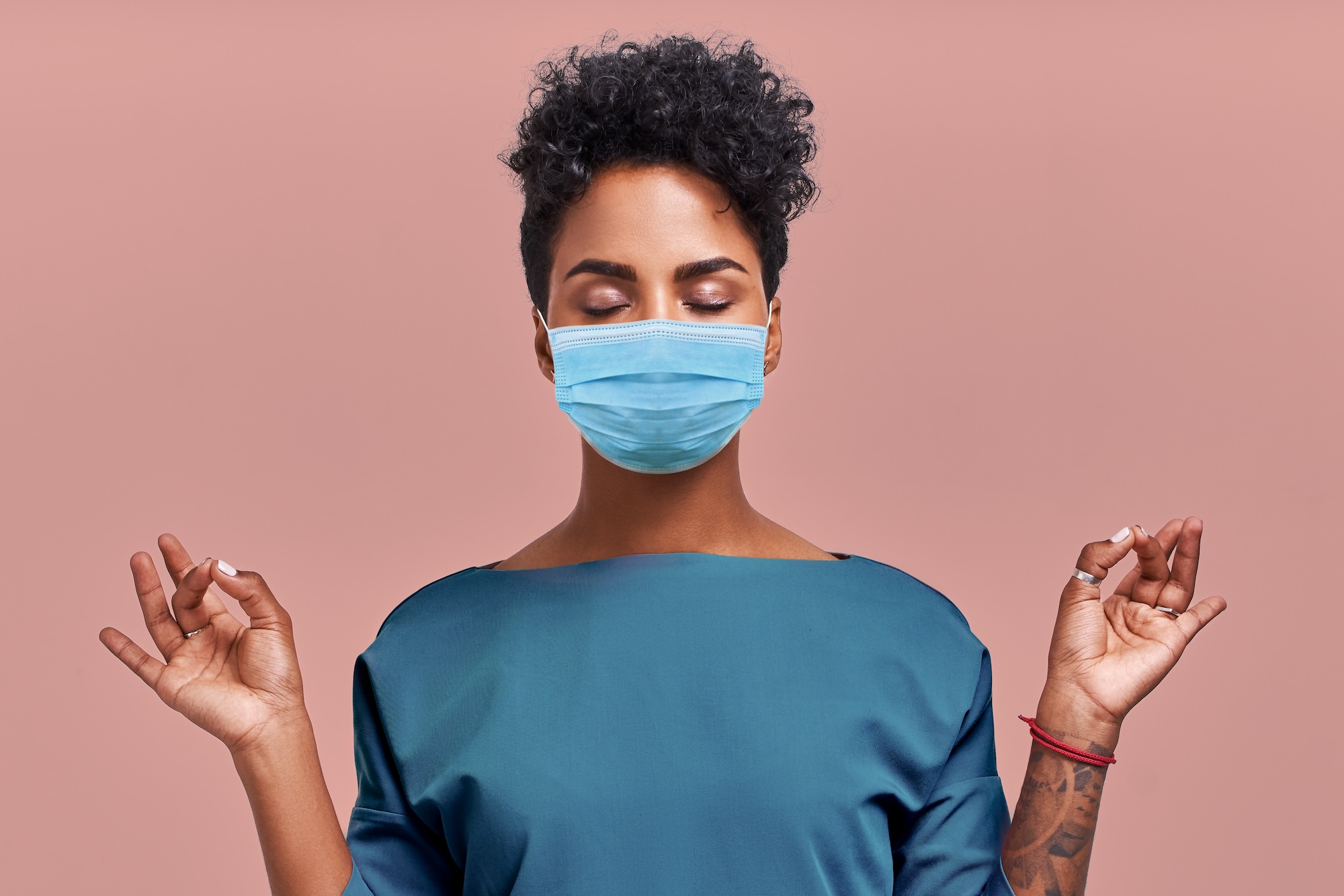 Person with curly hair wearing a blue face mask and teal shirt meditating with eyes closed on pink background.
