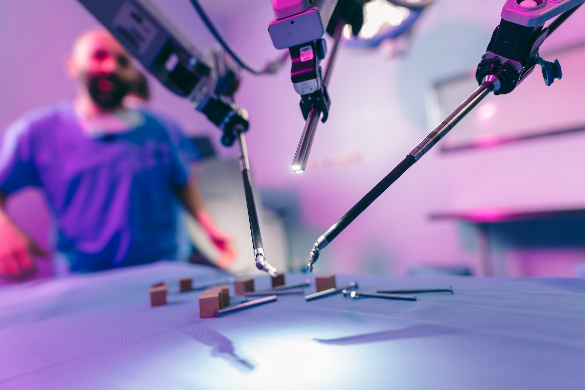 Robotic surgical arms operating on a table with medical tools and a surgeon blurred in the background.