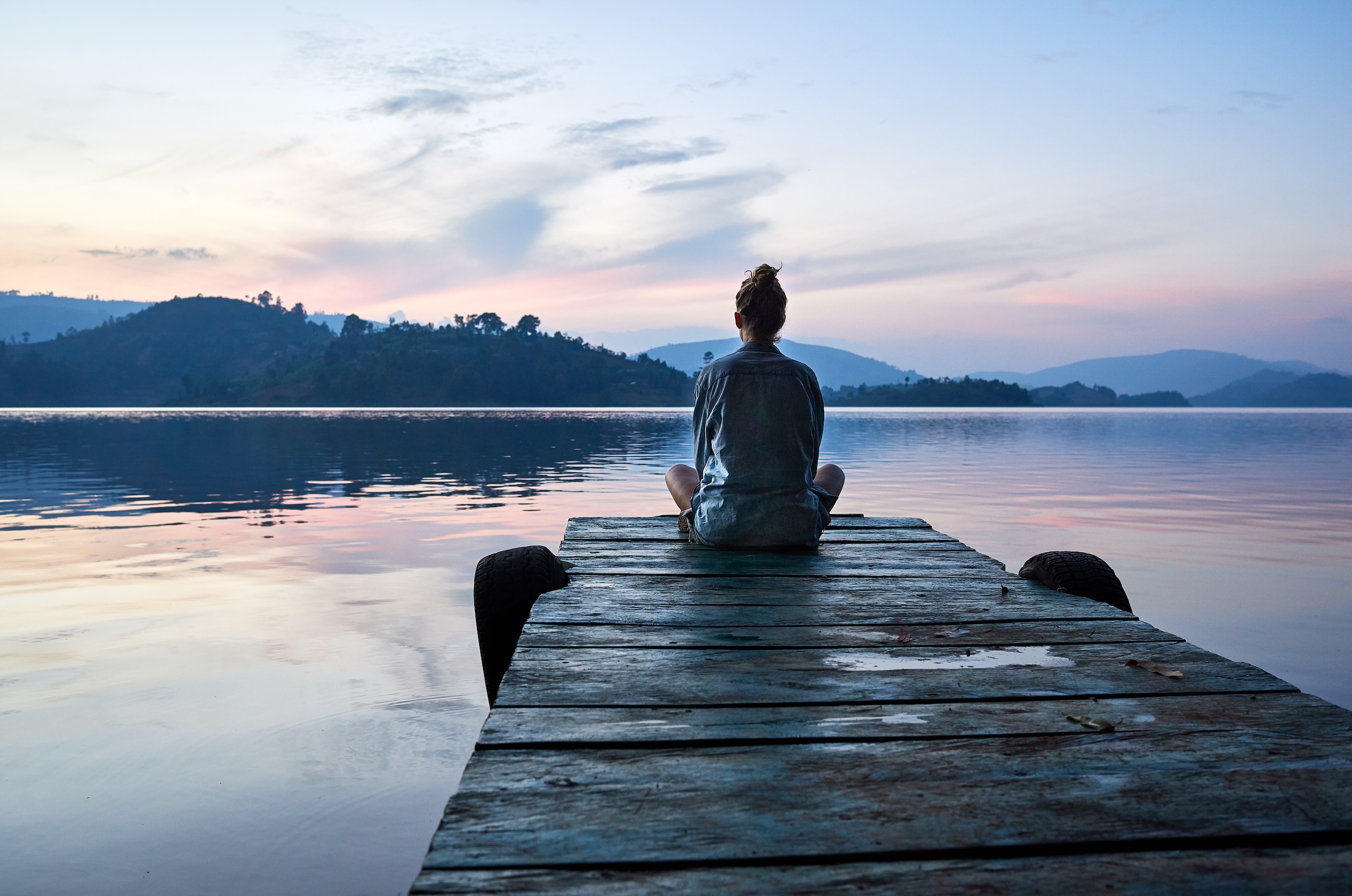 Person sitting alone on a wooden dock overlooking a calm lake at sunset with mountains in distance.