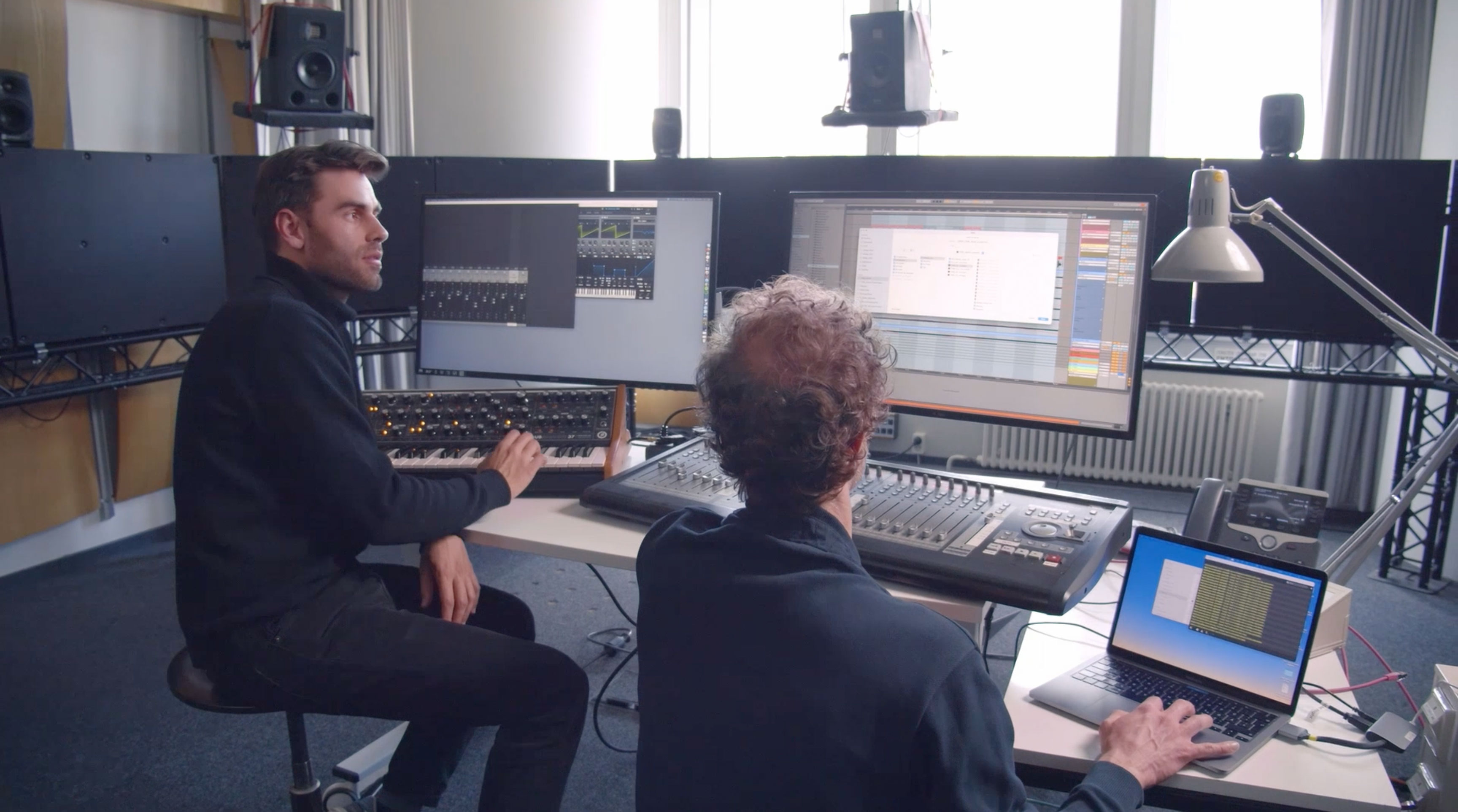Two men working in a music studio with mixing console, laptop, synthesizer, and multiple monitors.