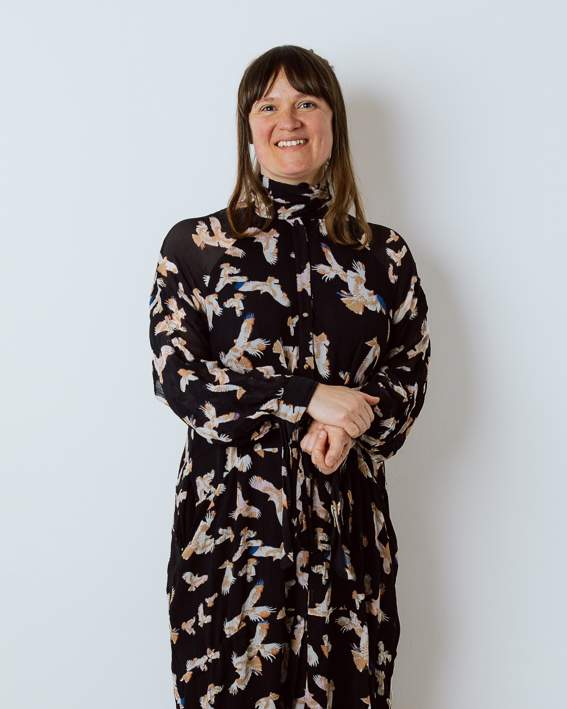 Smiling woman with shoulder-length hair wearing a black dress with bird prints against a white wall.