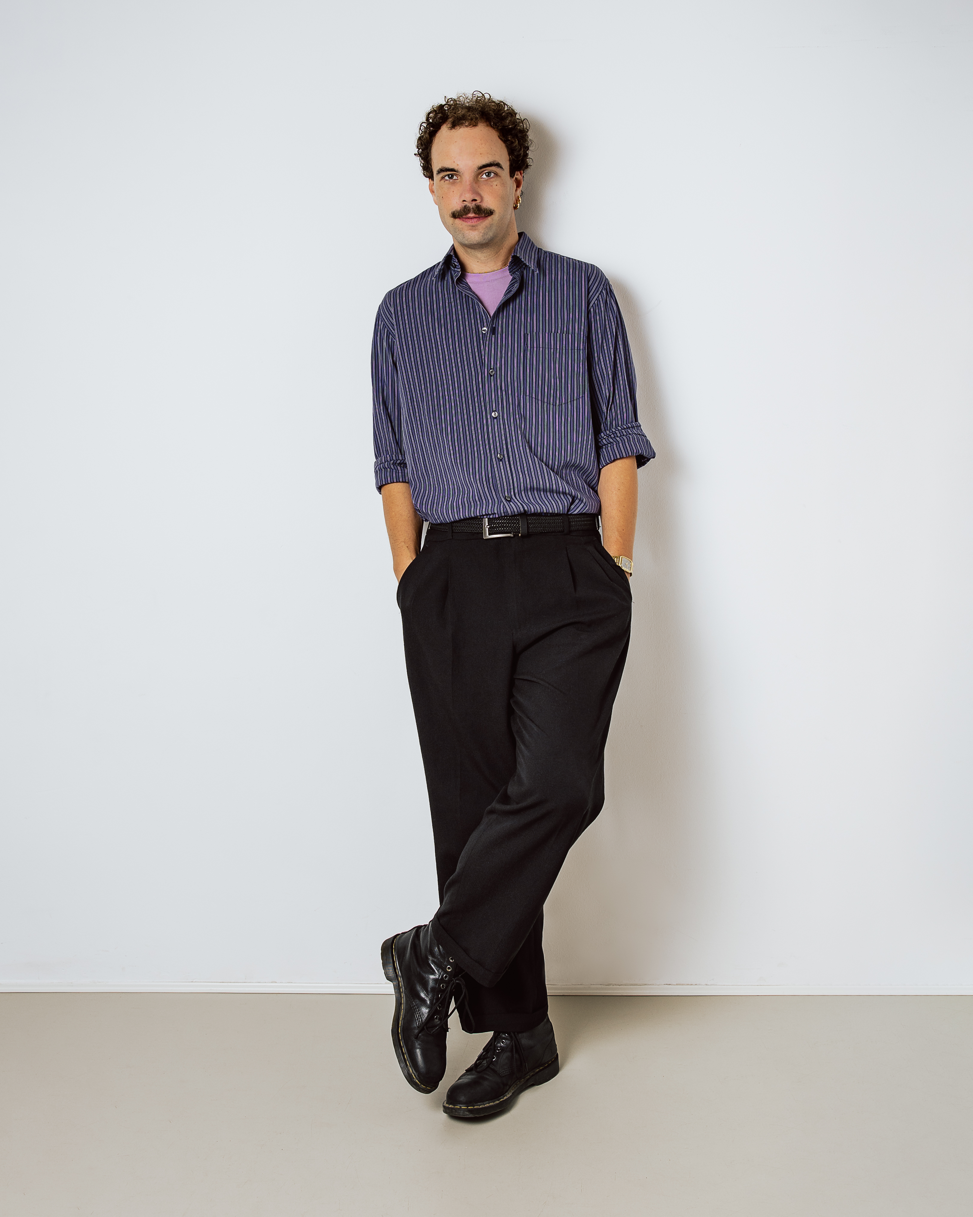 Man with curly hair and mustache wearing striped shirt and black pants, leaning against a white wall.