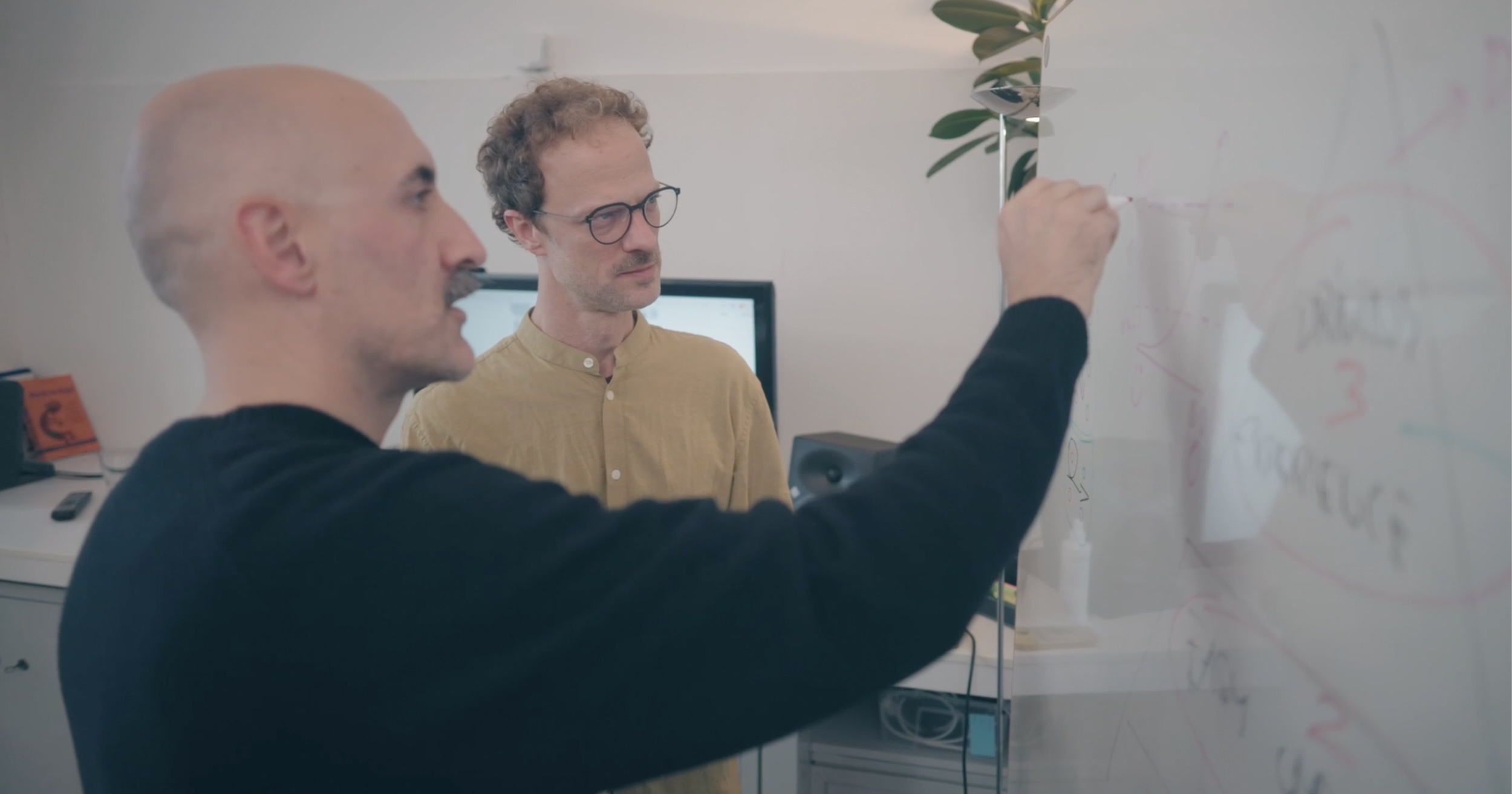 Two men, one writing on a transparent board, discussing ideas in an office setting.