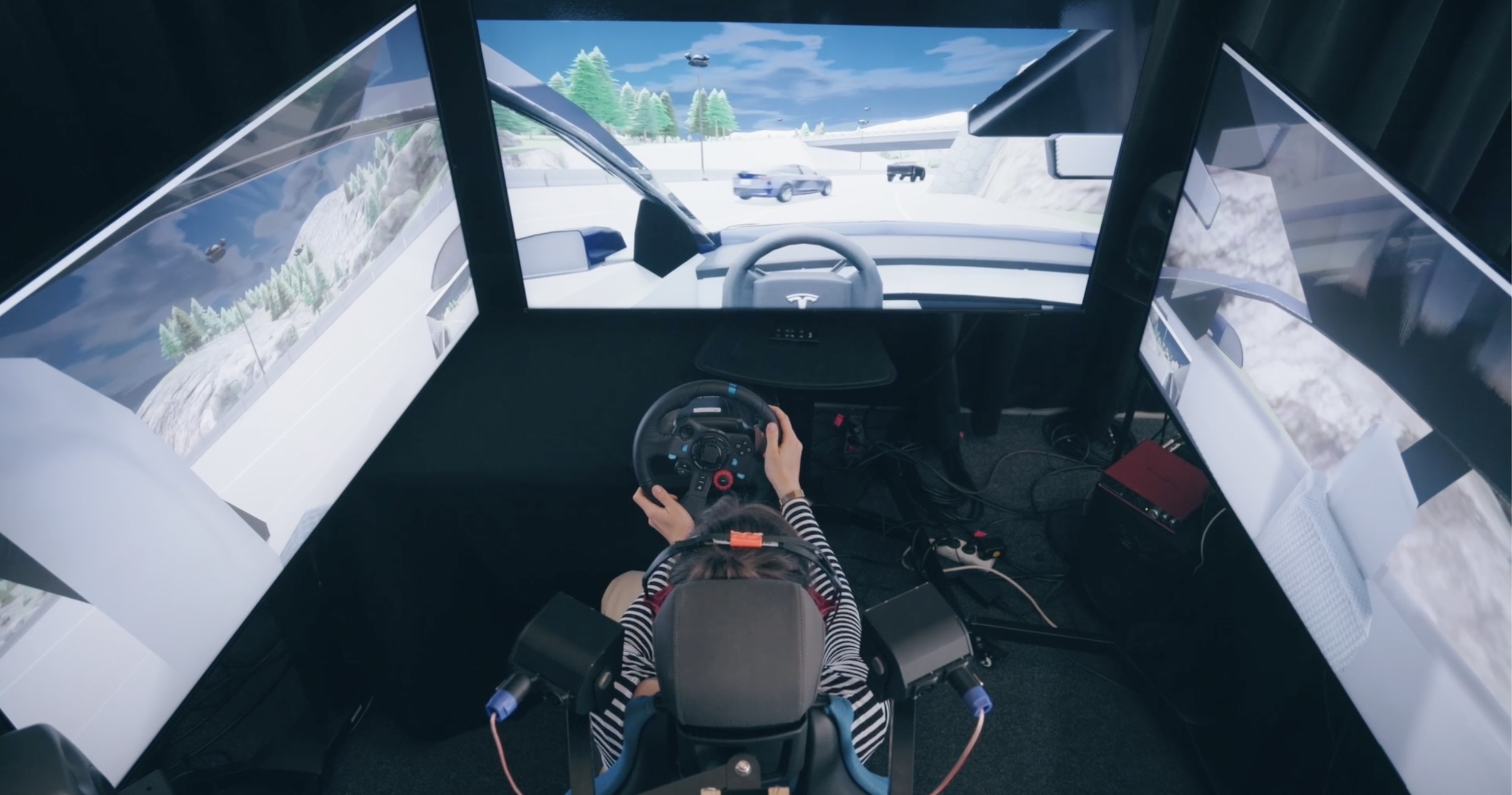 Person using a racing simulator with three screens and a steering wheel in a virtual driving environment.