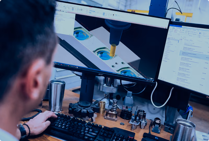 Person working on a computer with CAD software displaying 3D model of machinery parts and a CNC machine tool, with metal components and tools on the desk.