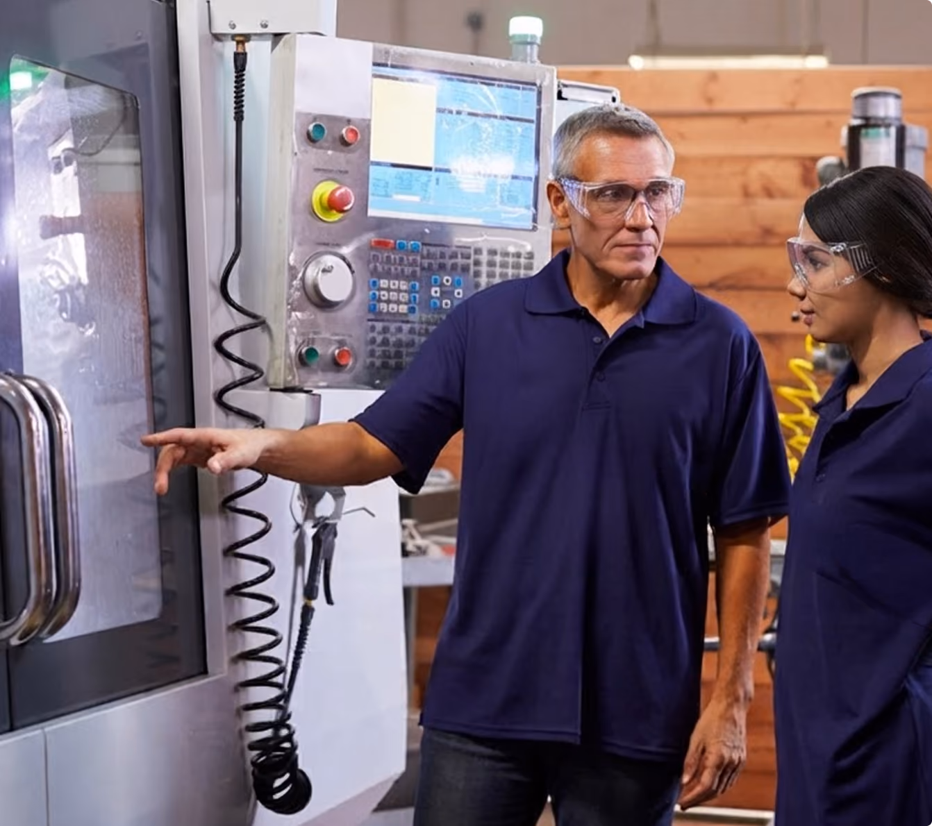 A man and woman wearing safety glasses and navy blue shirts stand near industrial machinery. The man points at a part of the machine while the woman attentively looks on, suggesting a discussion or training in a manufacturing or workshop setting.