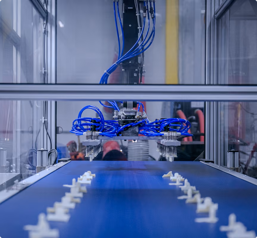 Automated robotic machinery with blue pneumatic tubes working above a conveyor belt transporting small white plastic components in a factory setting.