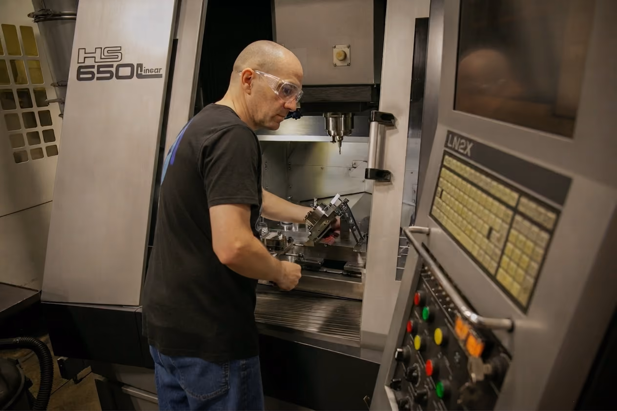 A man wearing safety glasses operates an HS 650 Linear CNC machine, adjusting metal parts inside the machining area in a workshop.