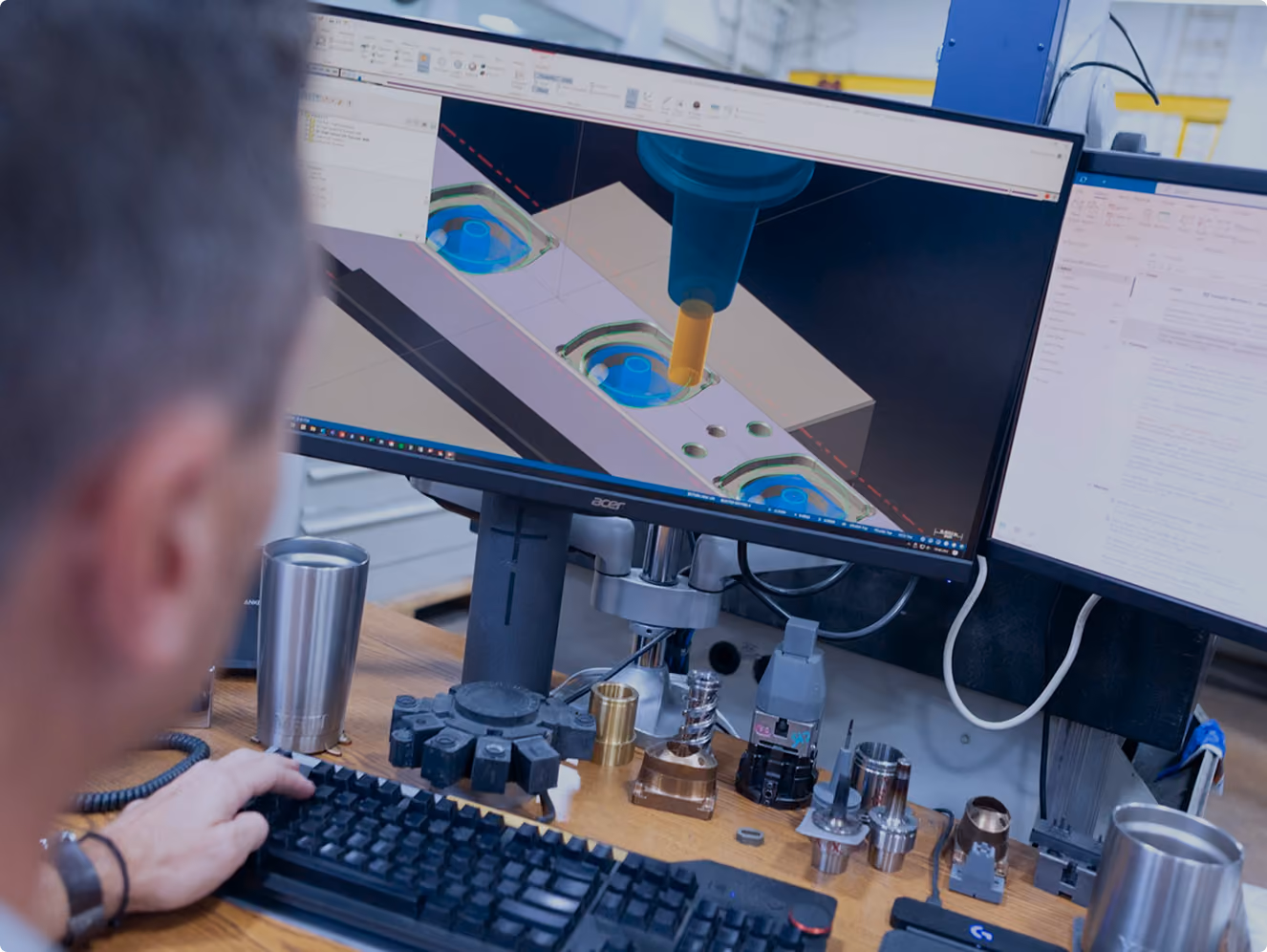 Person using CAD software on a computer to design or simulate a milling process for a mechanical part, with various metal parts and tools on the desk.