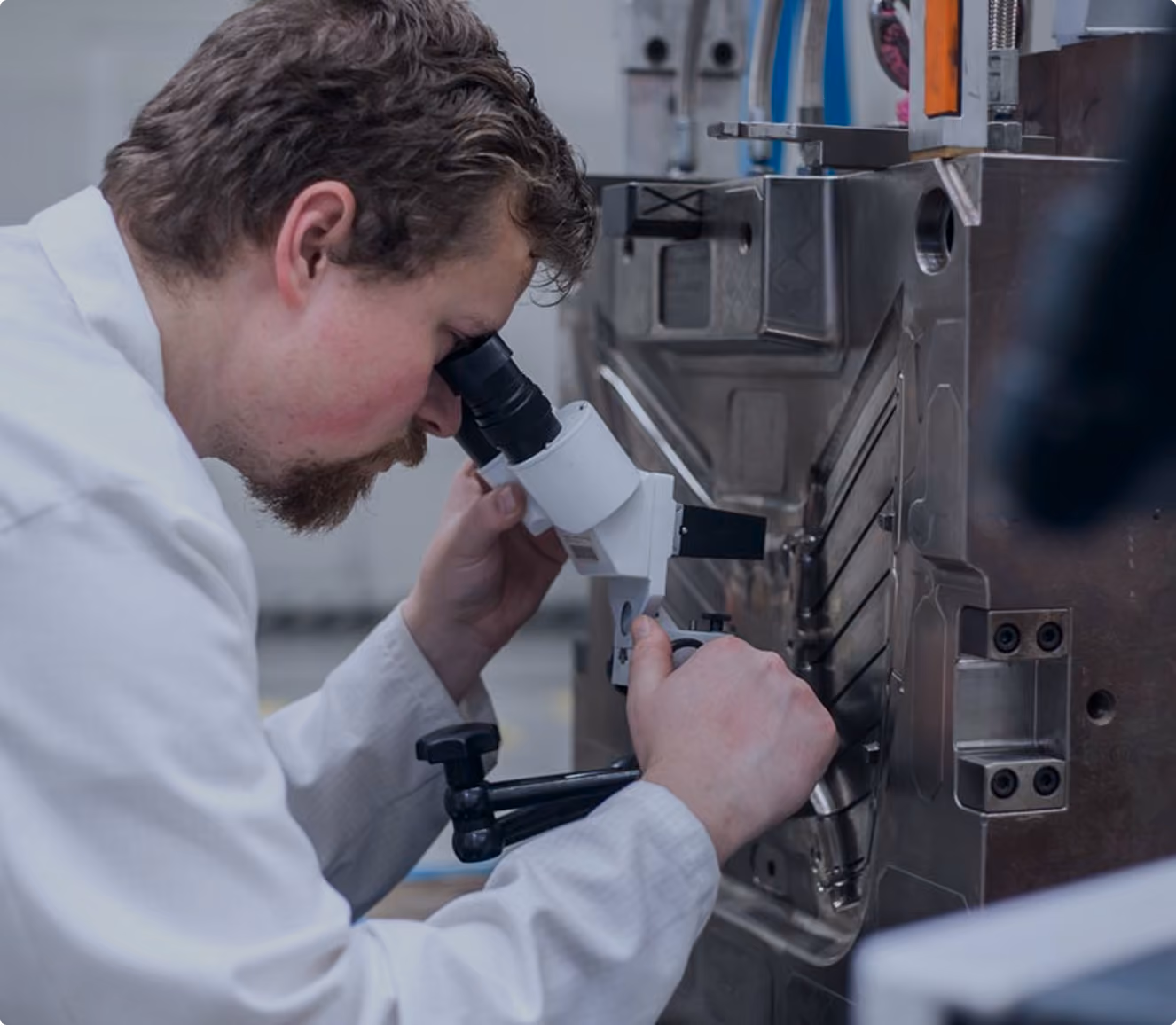 Man in a white lab coat closely examining machinery through a mounted monocular microscope in an industrial or laboratory setting.