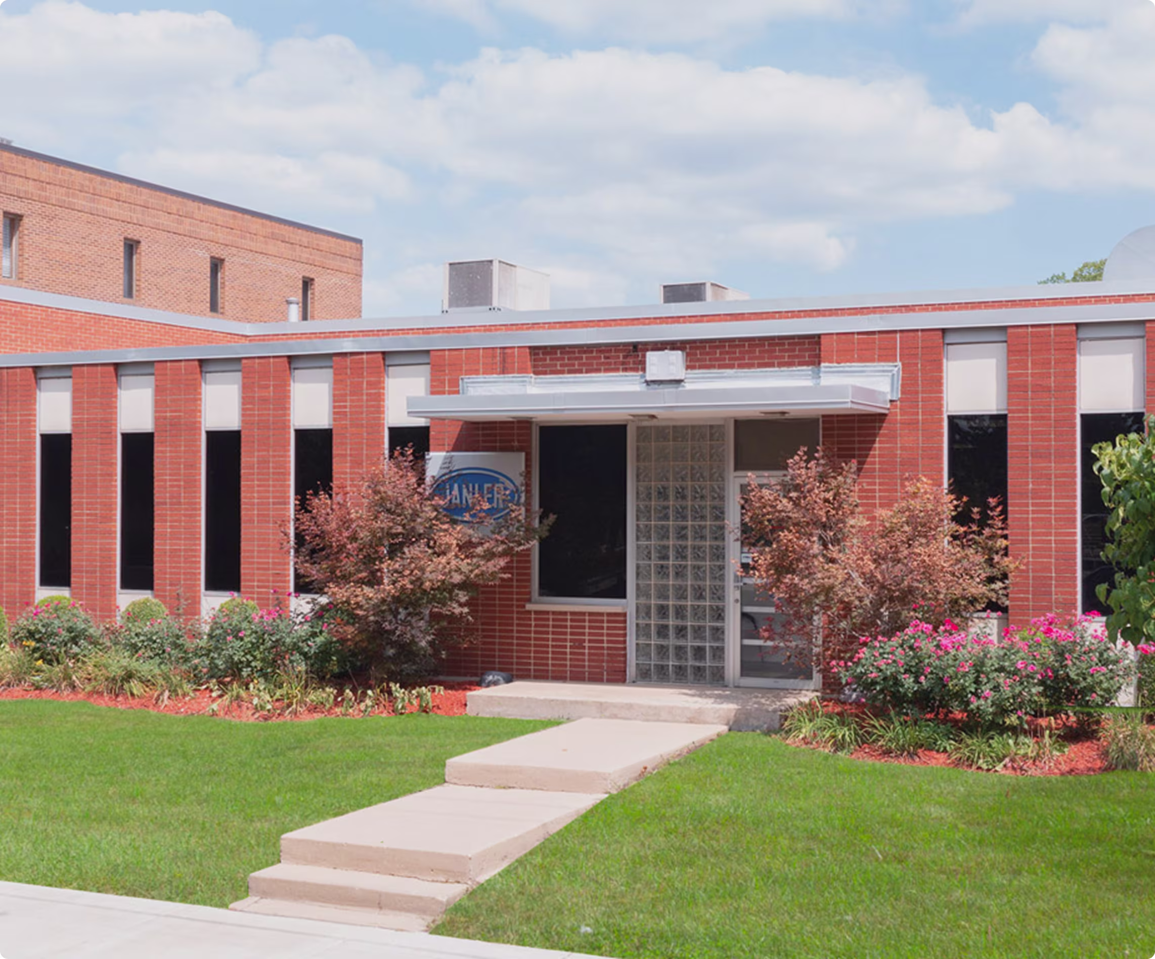 Single-story brick building with a glass block entrance door, black vertical windows, small overhang, surrounded by green lawn, bushes, and pink flowers under a partly cloudy sky.