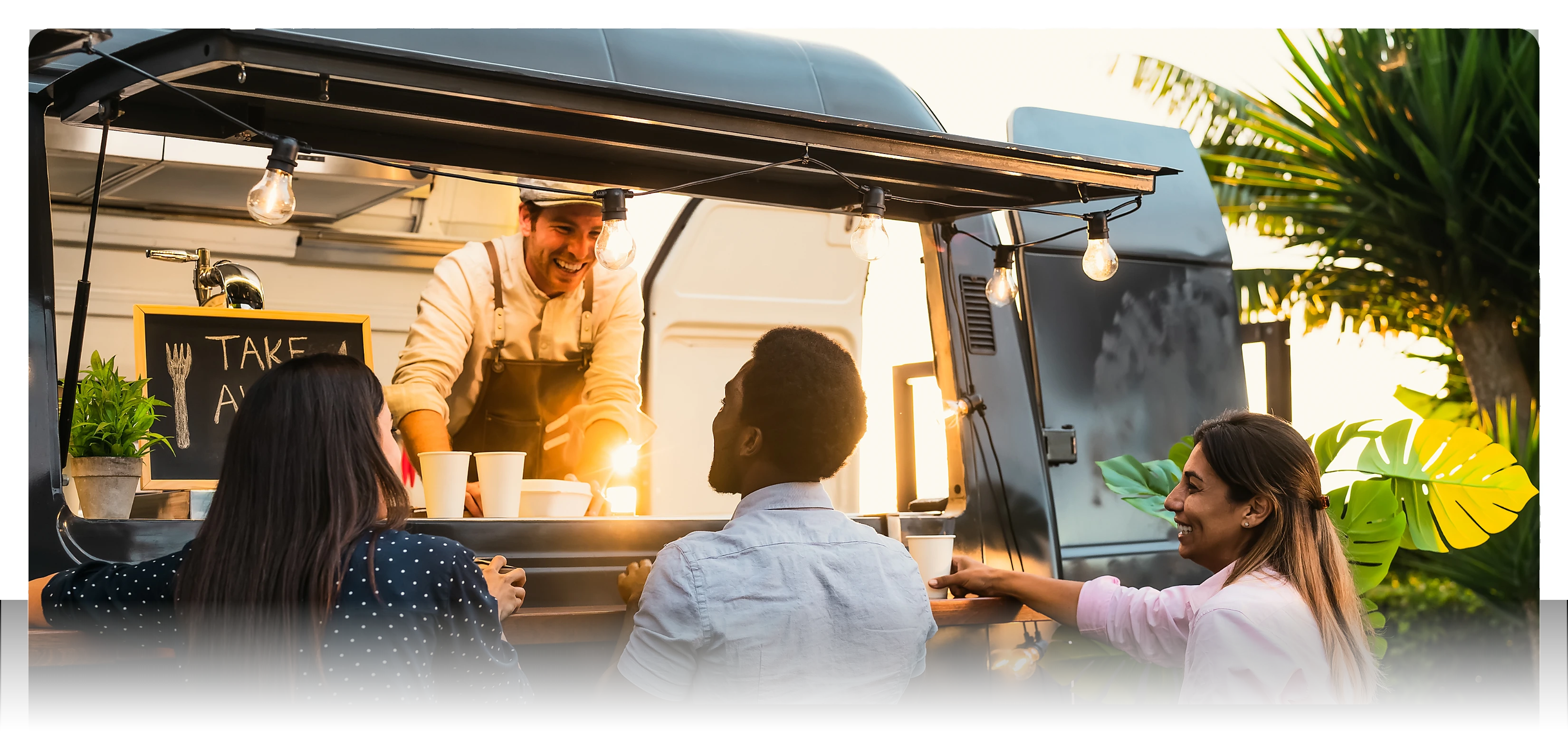 Two women in white uniforms serving food to customers at an outdoor food truck with illuminated menu signs.