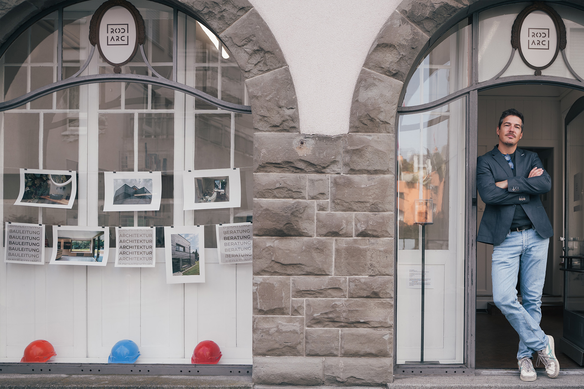 Mann lehnt an Türrahmen neben Fenster mit Architekturphotos und drei Helmen drinnen.