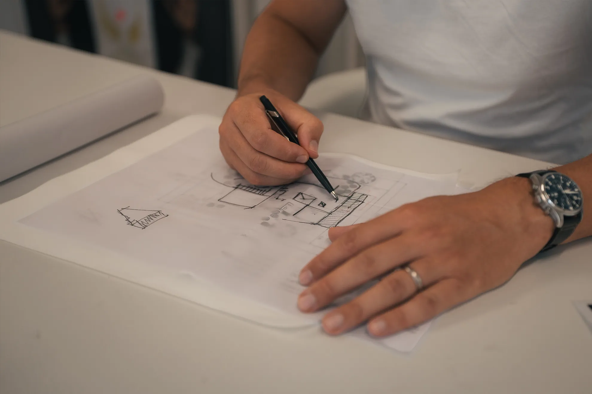 Person zeichnet einen Architekturplan mit einem Stift auf transparentem Papier am Tisch.
