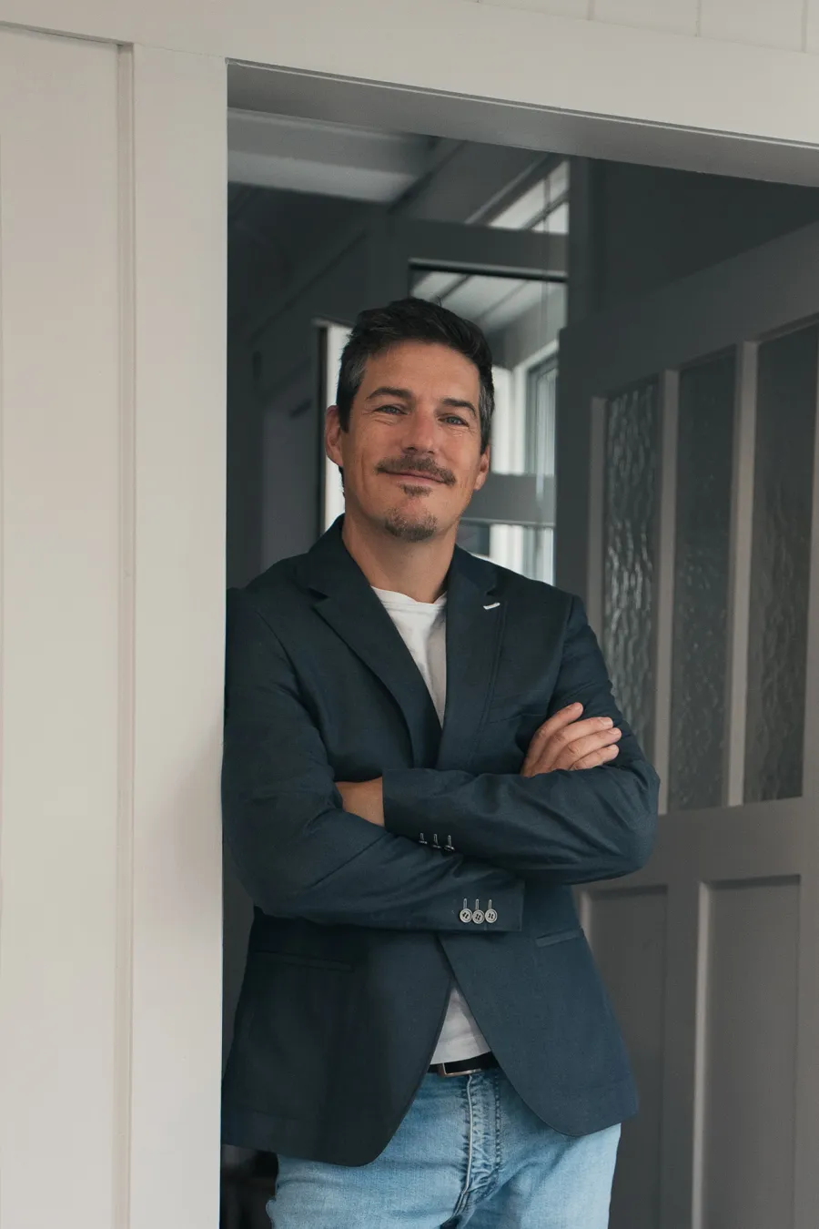 Ein Mann mit dunklem Sakko und Jeans steht mit verschränkten Armen in einer Türöffnung.
