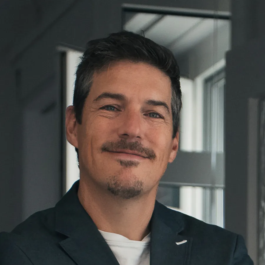 Mann mit dunklem Sakko und weißem T-Shirt lächelt vor Fenster in Büro.