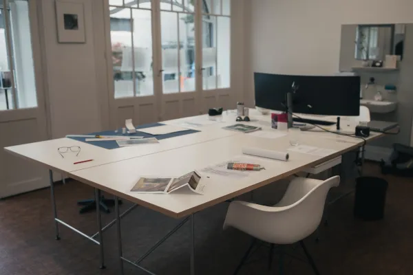 Ein moderner Büroraum mit großem Tisch, Stuhl, Papieren und Computer vor hellen Fenstern.