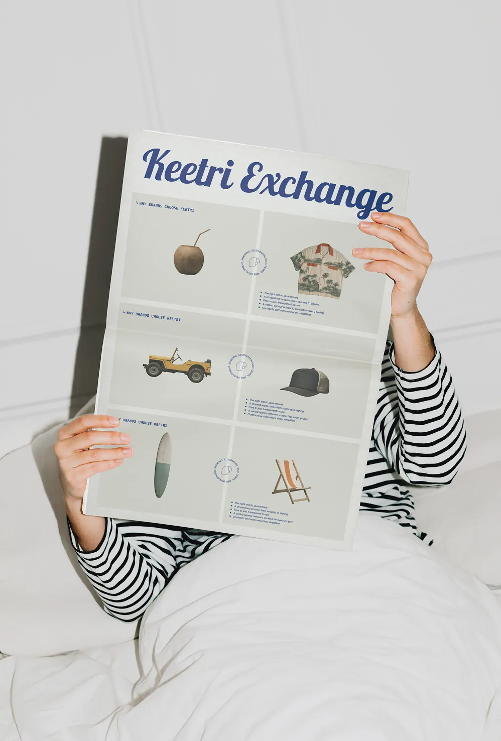 Person holding a Keetri Exchange catalog page in bed, illustrated with tropical items like a coconut, jeep, and surfboard