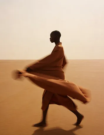Silhouette of a person walking barefoot on desert-like sand, wearing a flowing brown garment caught in motion against a plain beige sky.