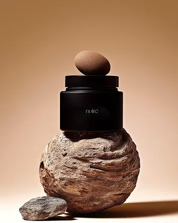 A minimalist composition featuring a black container labeled 'noir.' balanced on a textured, round stone with a brown egg placed on top, set against a smooth beige background.