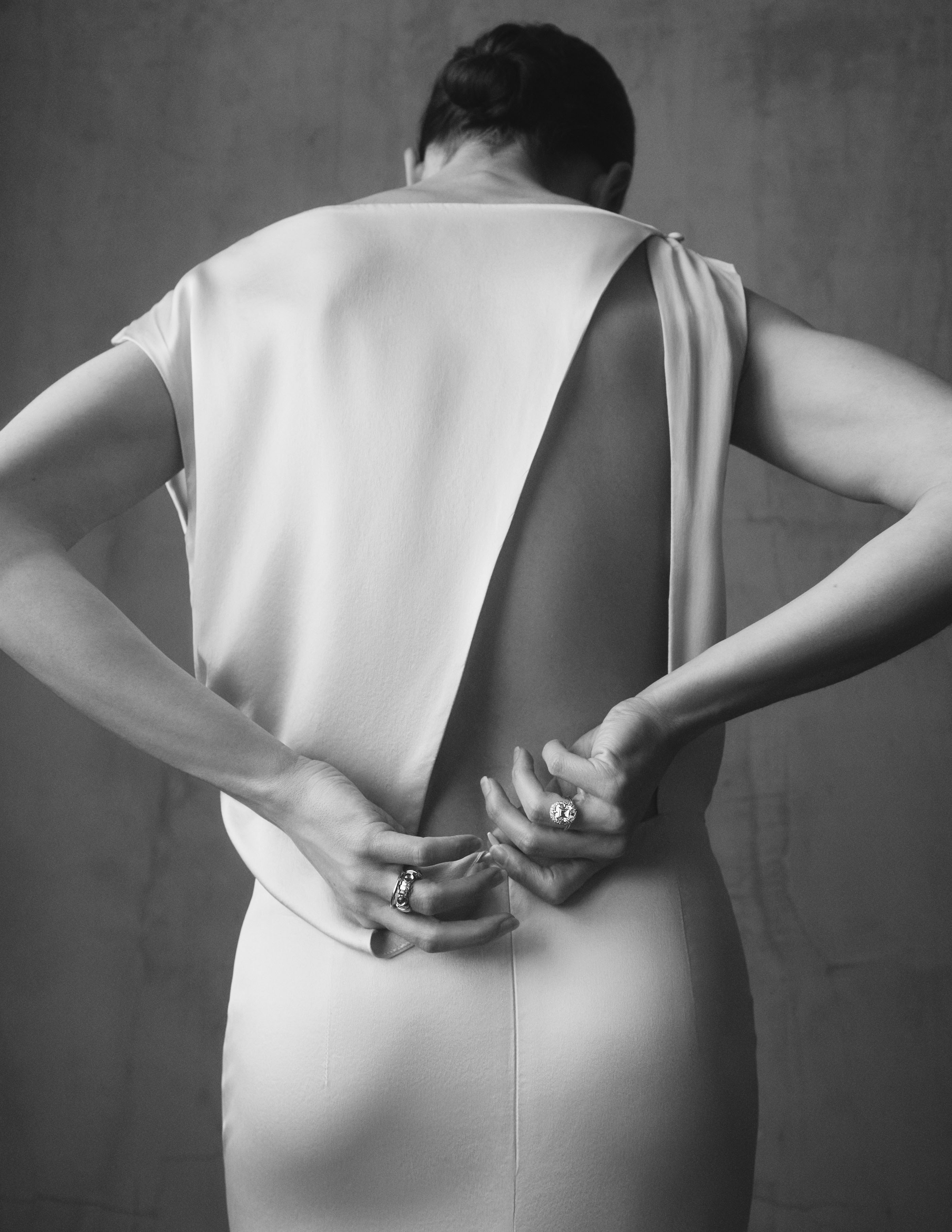 Black and white photo of a woman in a backless dress fastening it behind her back, wearing two rings.