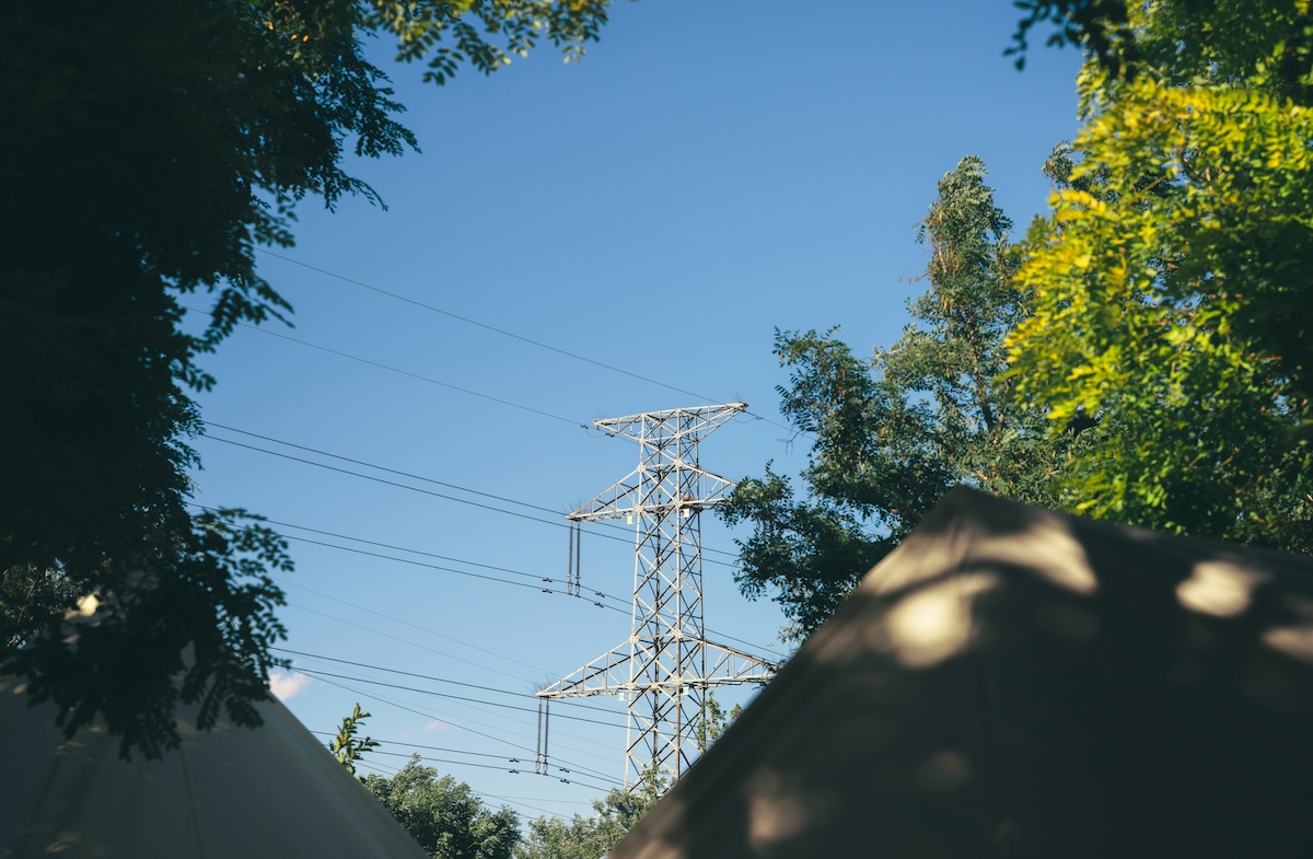 Metal electricity transmission tower with power lines among green trees under a clear blue sky.
