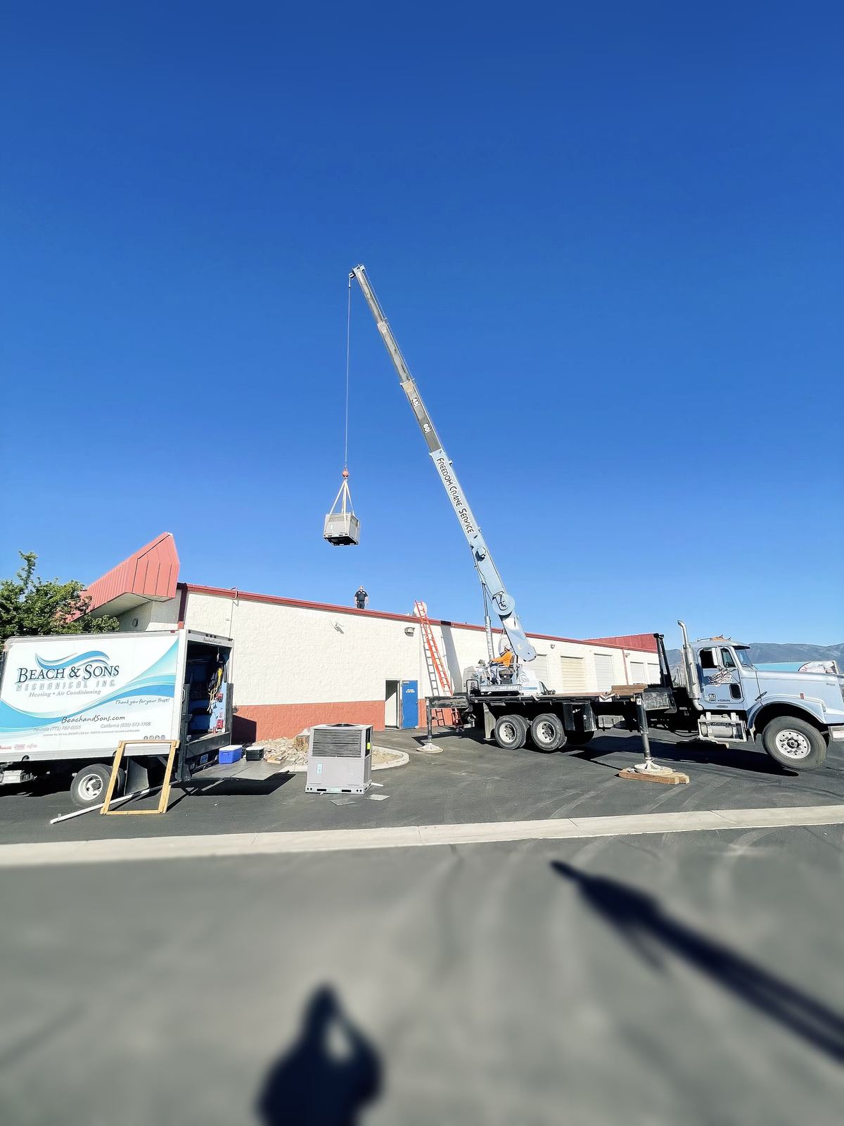Crane lifting HVAC equipment at a commercial job site with a Beach and Sons truck below