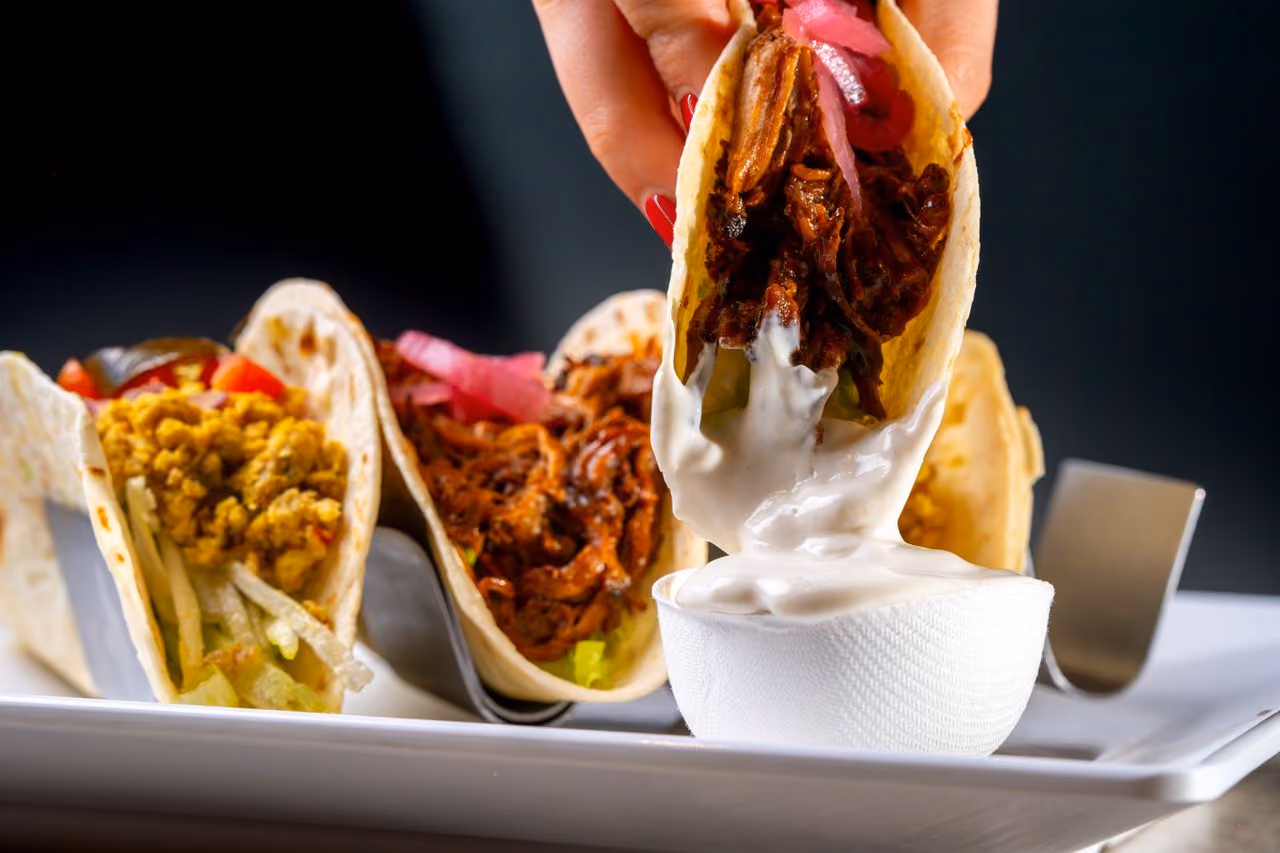 Hand holding meat taco dipped in white sauce with other tacos on a plate in background.