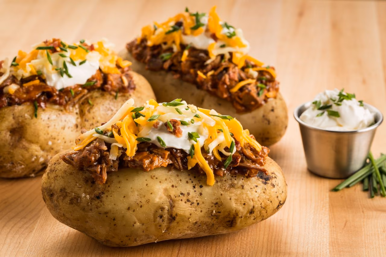 Three baked potatoes topped with shredded meat, cheddar cheese, sour cream, and chopped chives on wood table.