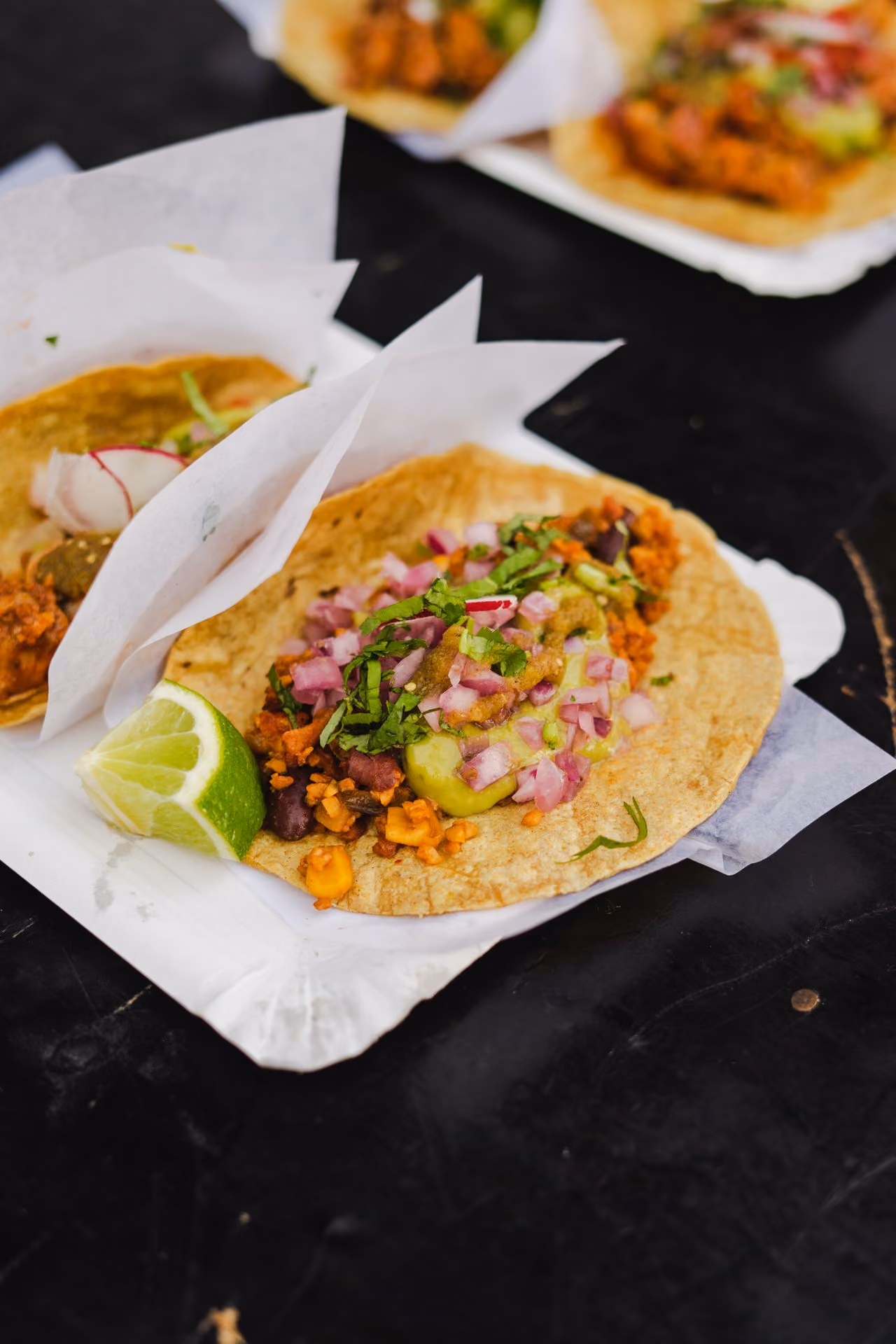 Close-up of tortilla taco with seasoned filling, avocado sauce, diced onions, cilantro, and a lime wedge on the side.