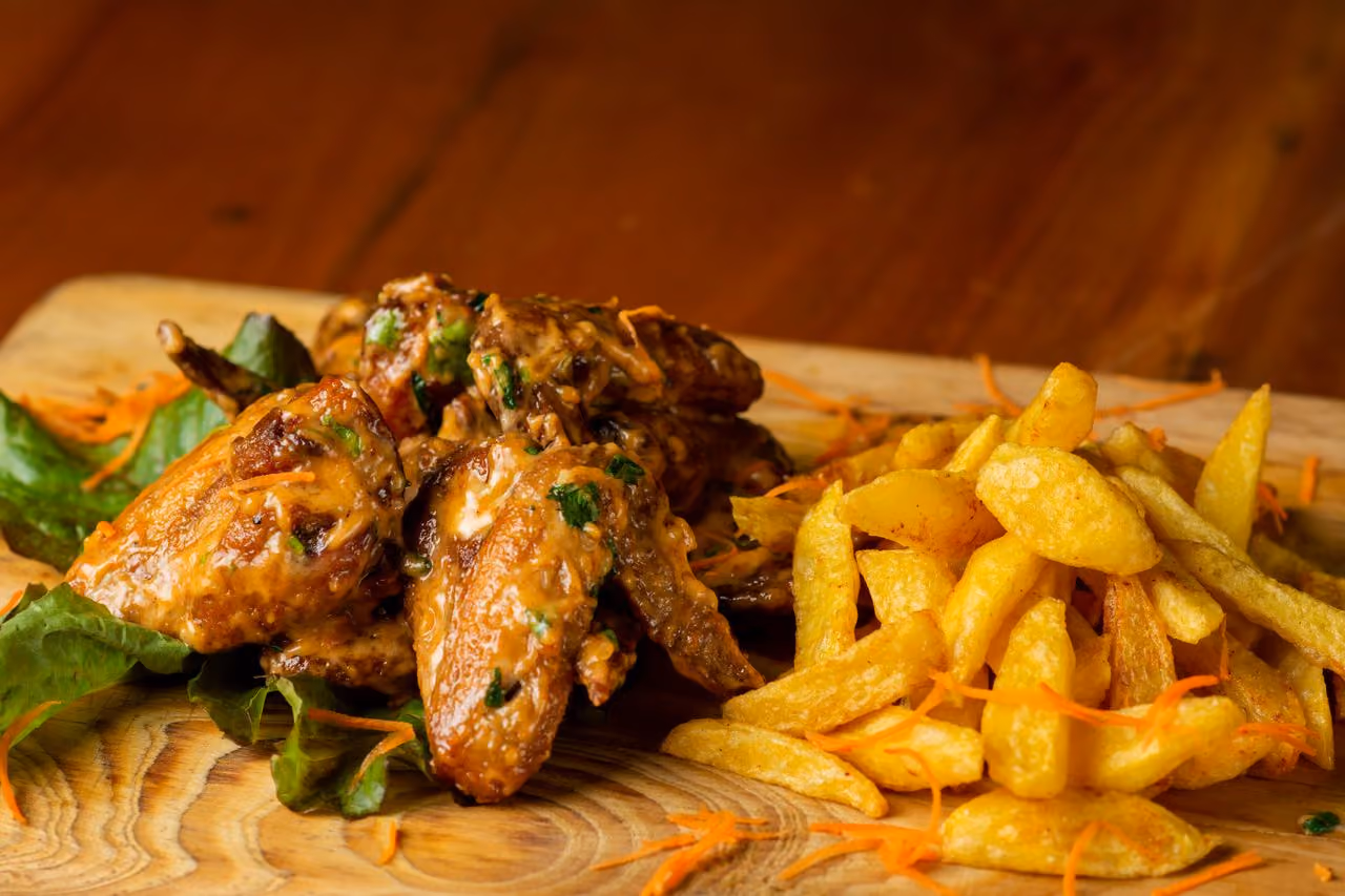 Plate of glazed chicken wings with herbs and crispy golden French fries on a wooden board.