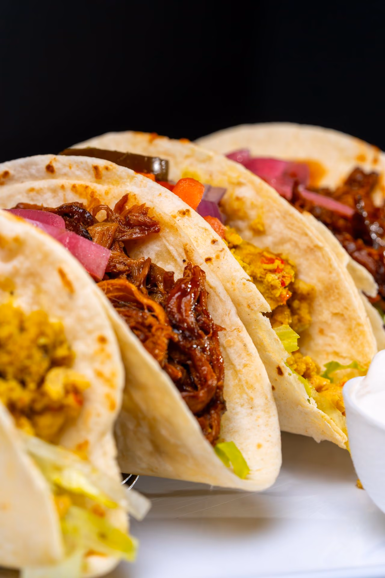 Close-up of three soft tacos with shredded meat, diced vegetables, and lettuce on a white plate with sour cream.