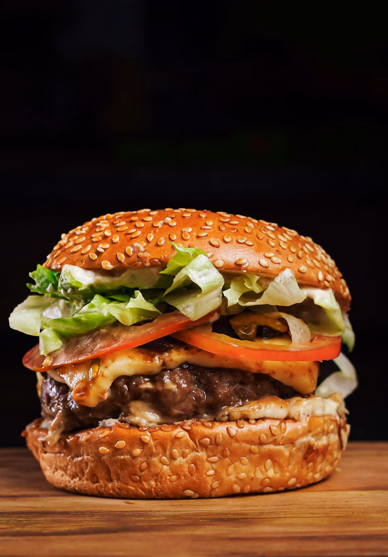 Cheeseburger with lettuce, tomato, and sesame seed bun on a wooden surface with black background.