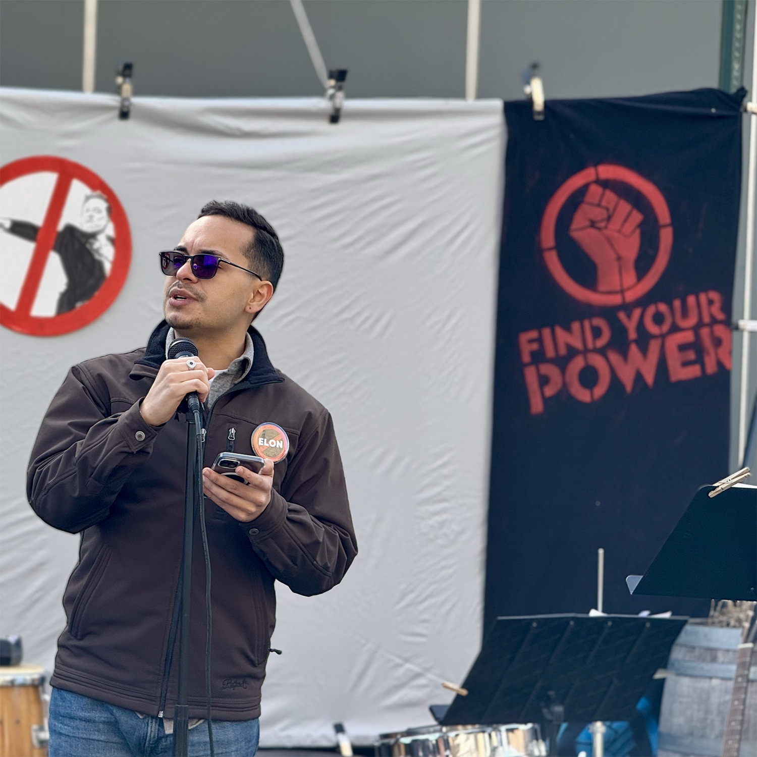Man wearing sunglasses and a brown jacket with an Elon badge speaking into a microphone while holding a phone, standing in front of a backdrop with a red fist and the words 'Find your power' and a no-kick symbol.