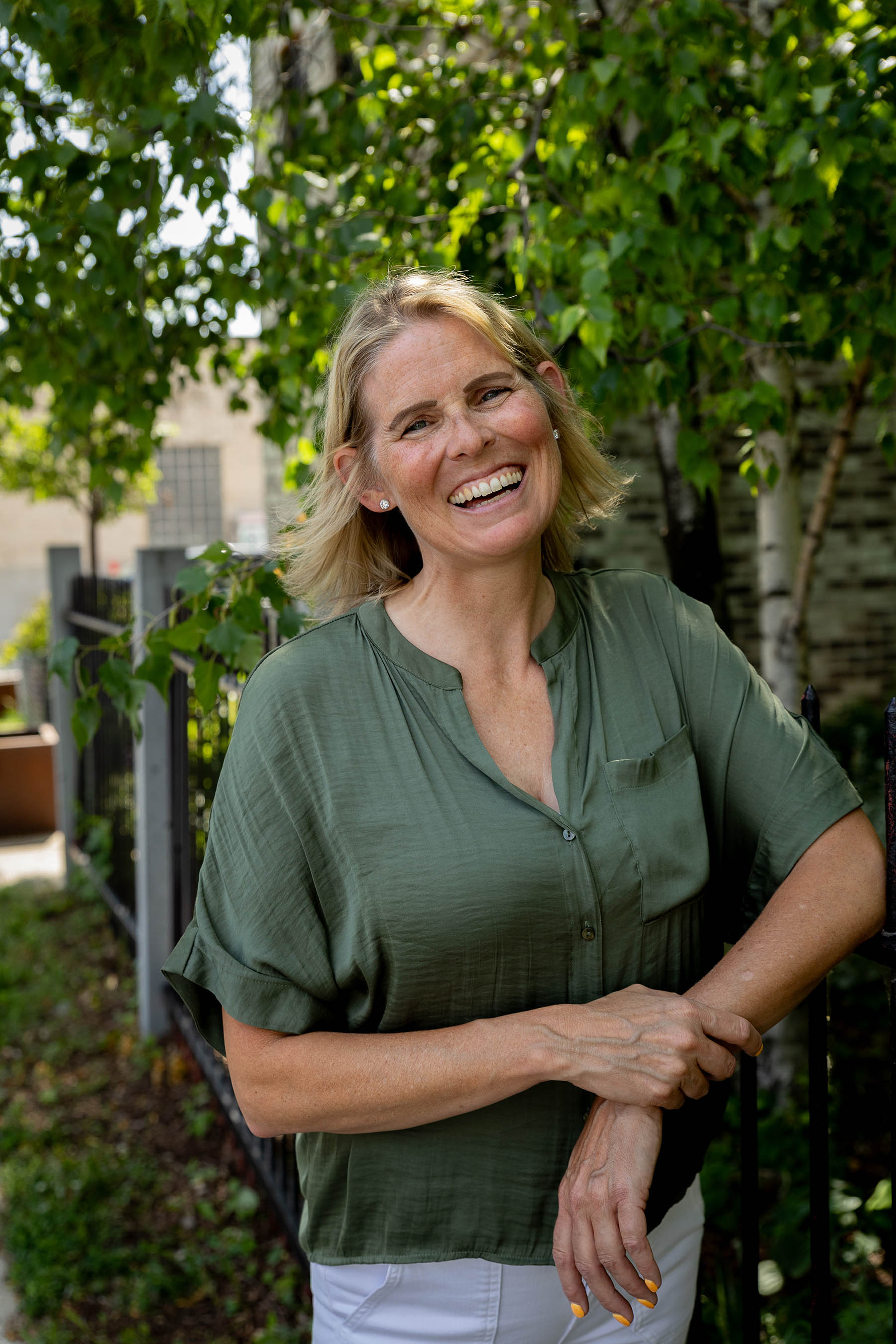 Smiling woman with short blonde hair wearing a green button-up blouse and white pants, standing outdoors by a black metal fence with green leafy trees in the background.