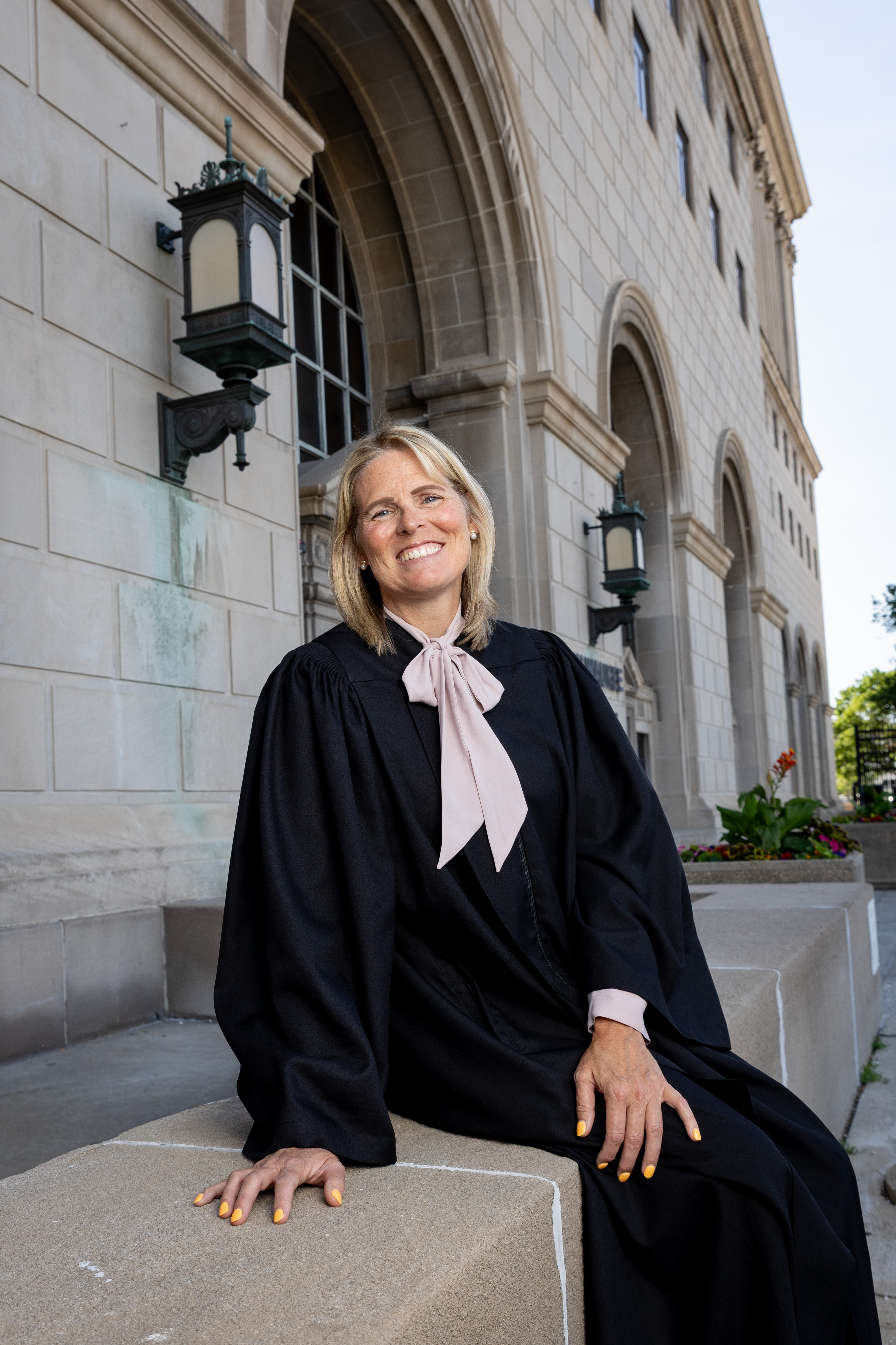 Smiling woman in a black judge's robe with a pale pink necktie sits on a stone bench outside a large stone building with arched windows and decorative lamps.