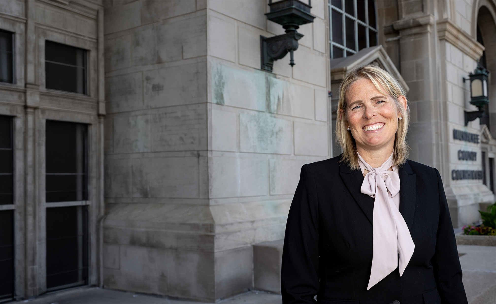 A smiling blonde woman wearing a black blazer and light pink bow tie blouse stands outside a building with the sign 'Milwaukee County Courthouse'.