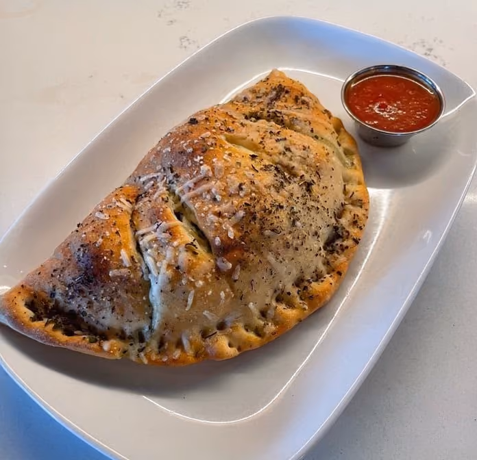Baked calzone sprinkled with cheese and herbs served with a cup of marinara sauce on a white plate.