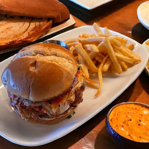 Chicken sandwich with melted cheese and fries served on a white plate with dipping sauce nearby.