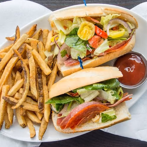 Plate with a deli sandwich stuffed with vegetables and meats, served with seasoned French fries and ketchup.