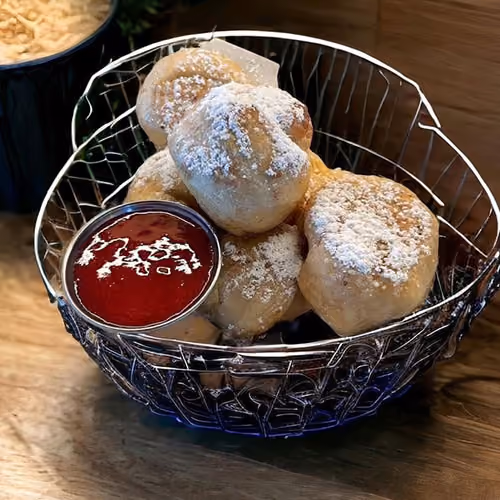 Basket of round fried pastries dusted with powdered sugar served with a cup of red dipping sauce.