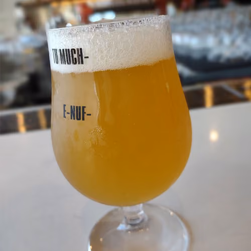 Tulip glass filled with hazy golden beer with foam head, text reads 'TU MUCH E-NUF'.